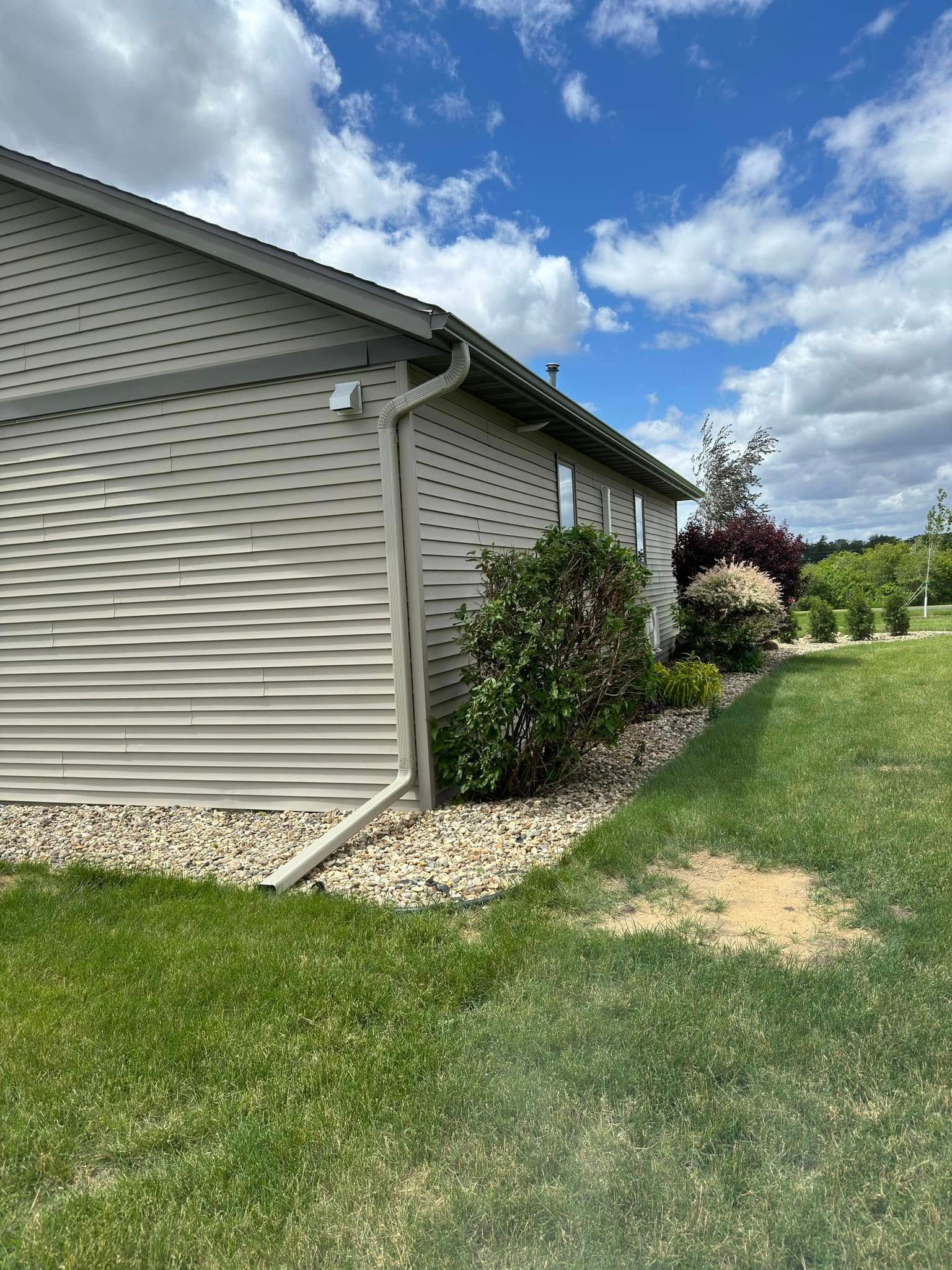 Beige house exterior with a gutter, green lawn, rock bed, and cloudy sky.