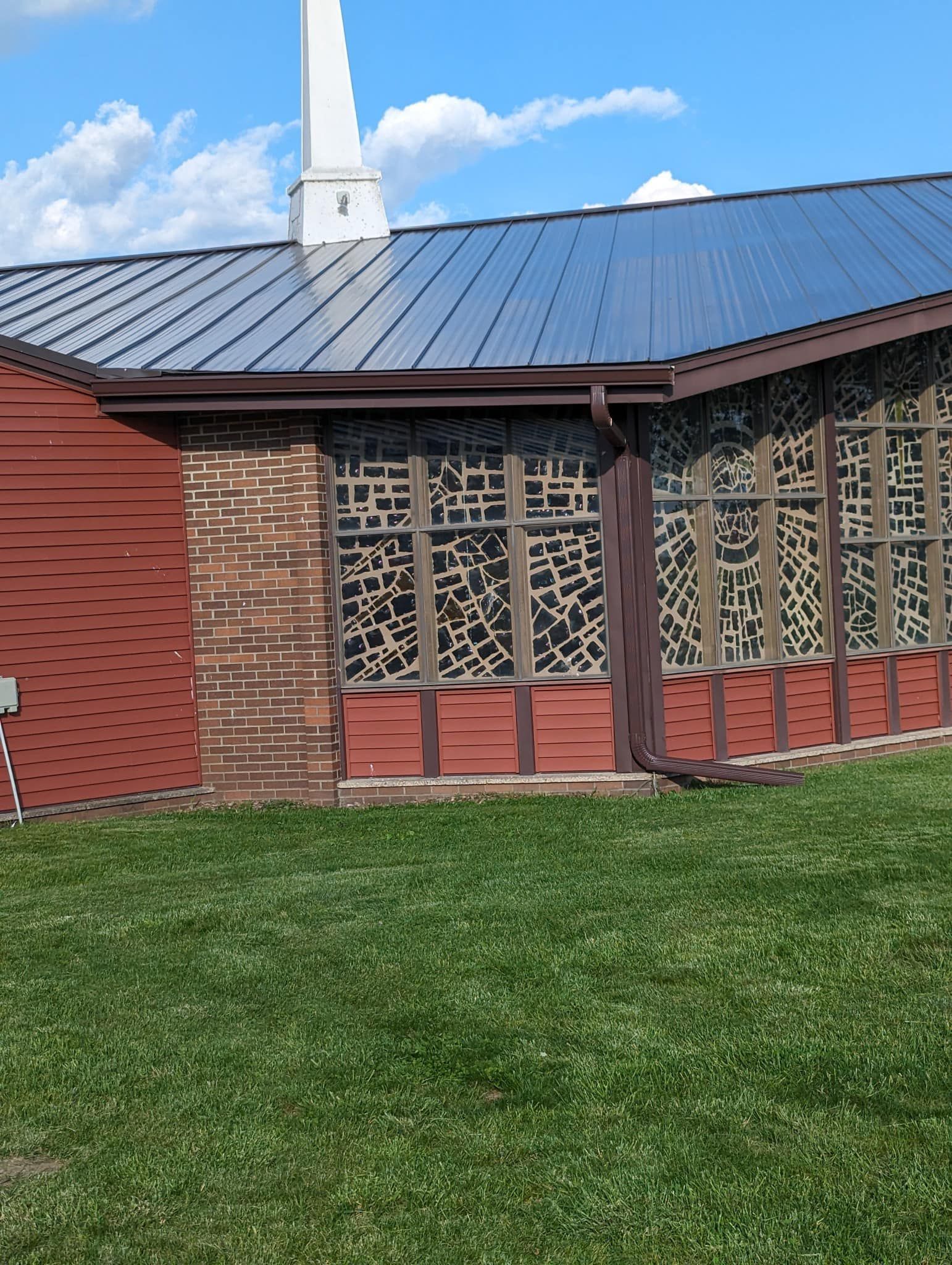 Red building with blue roof, steeple, and stained glass windows on a grassy lawn under a blue sky.