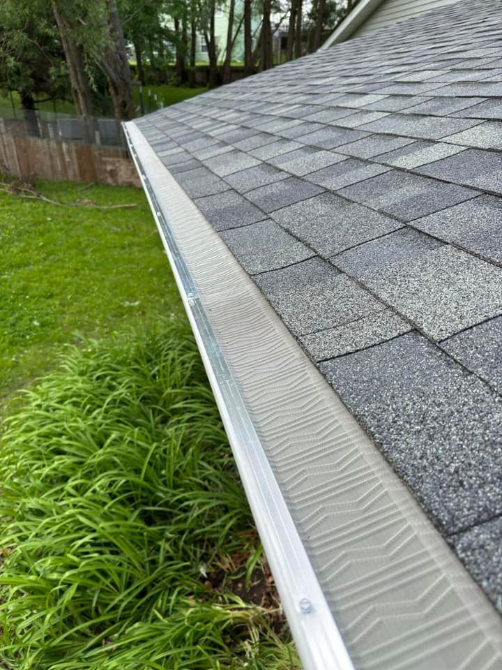 Gutter guard installed on a gray shingled roof with green grass and foliage in the background.