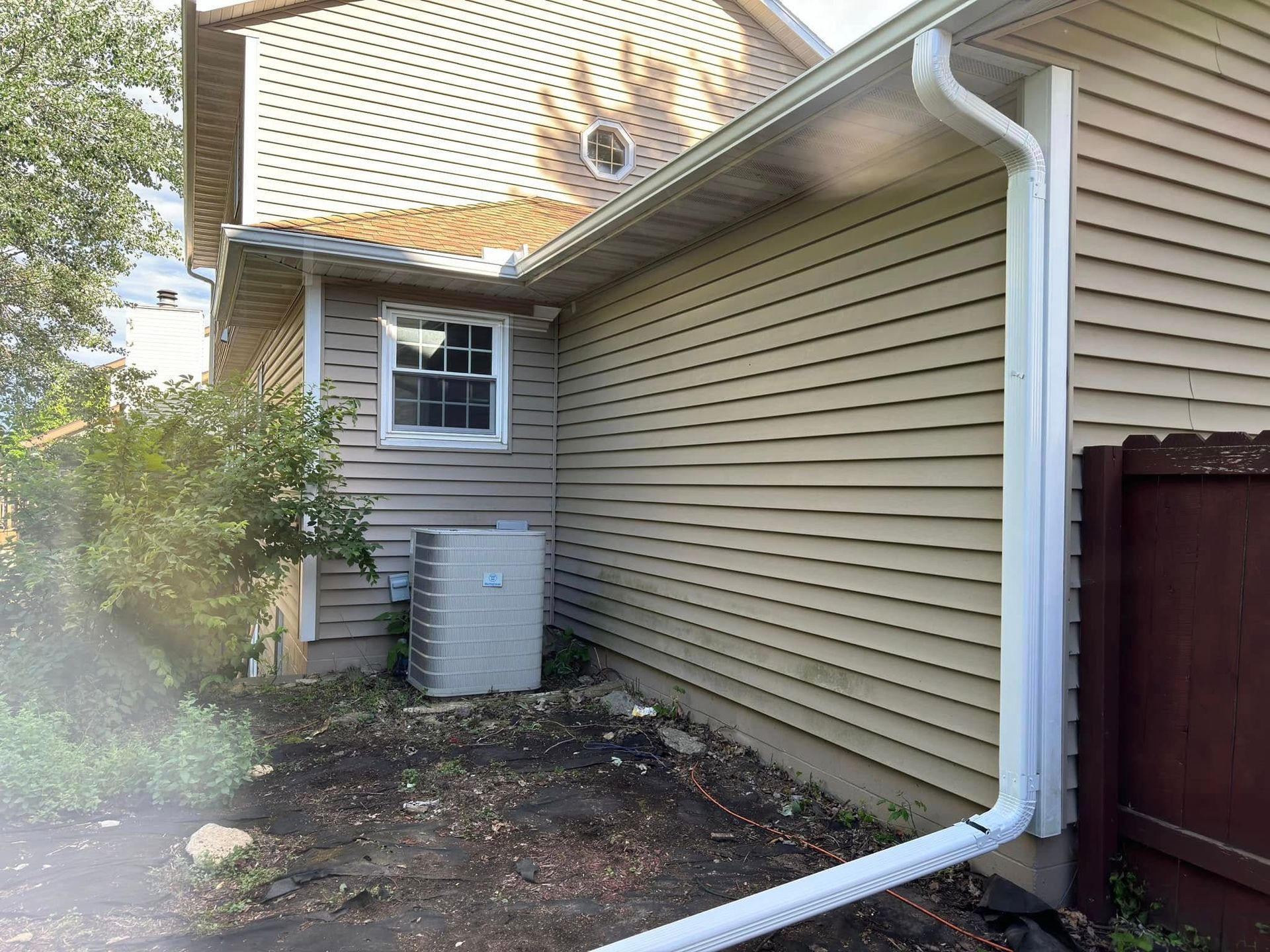 Side of a house with beige siding, white gutters, and an AC unit. A small window and a brown fence are visible.