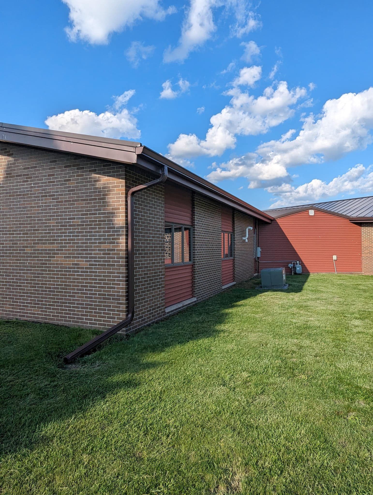 Brick building with brown roof, gutter, and green lawn under a blue sky with clouds.