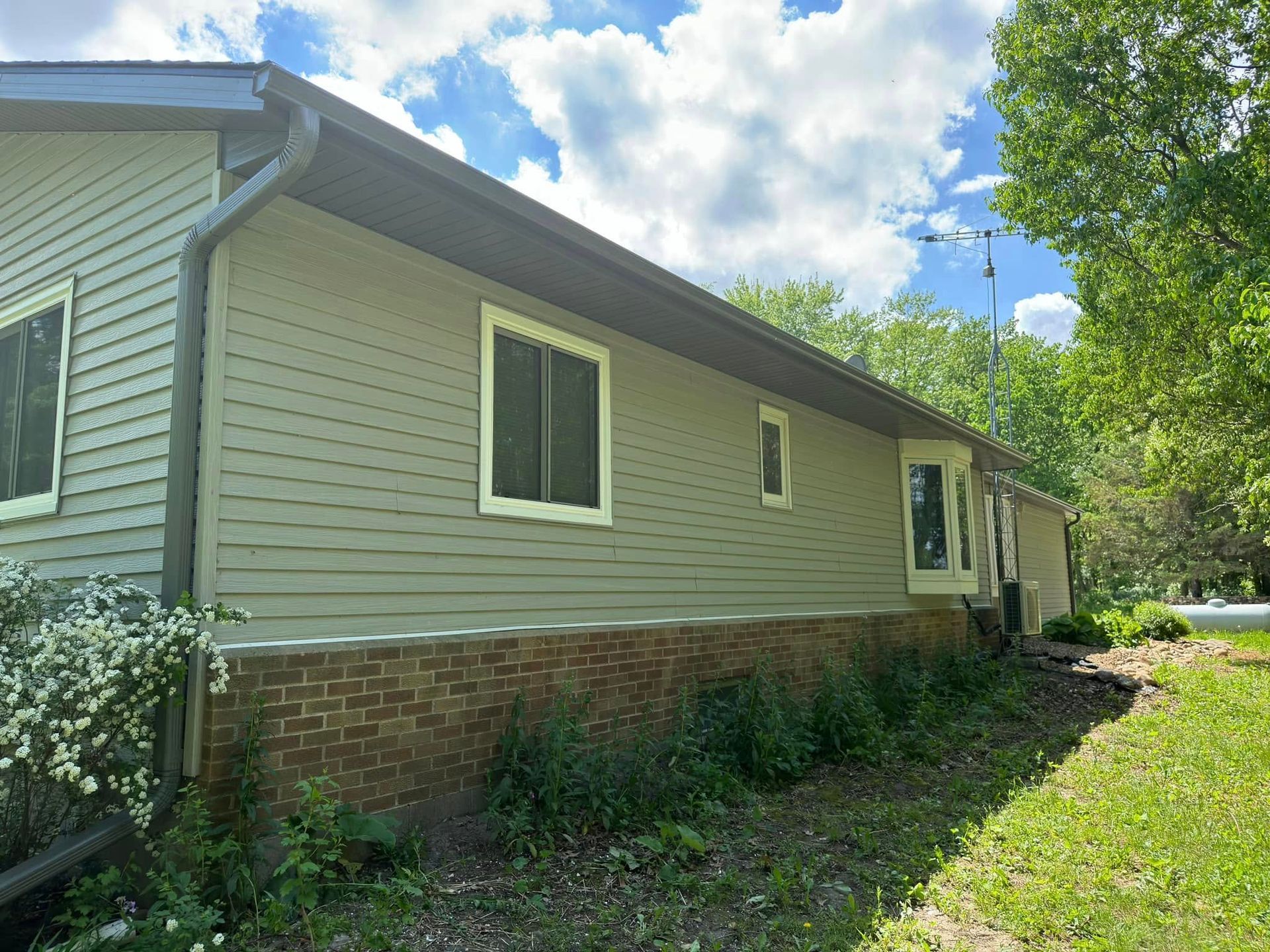 Exterior view of a house with green siding, brick base, and windows. Overcast sky with trees.