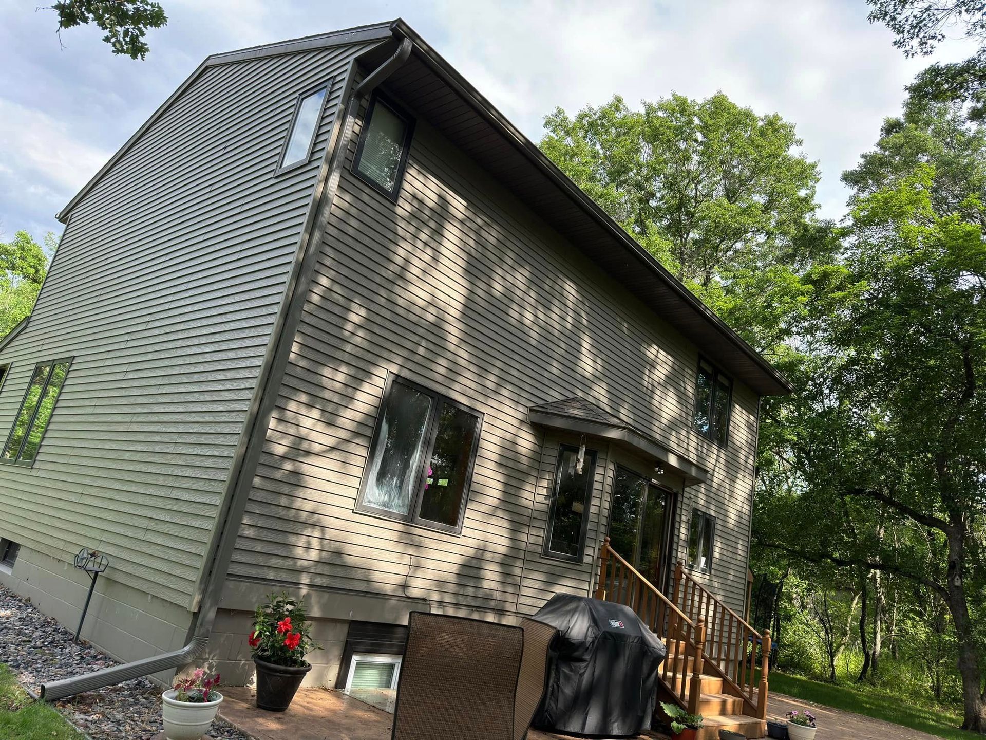Two-story house with green siding, brown trim, and wooden steps. Trees and cloudy sky in background.