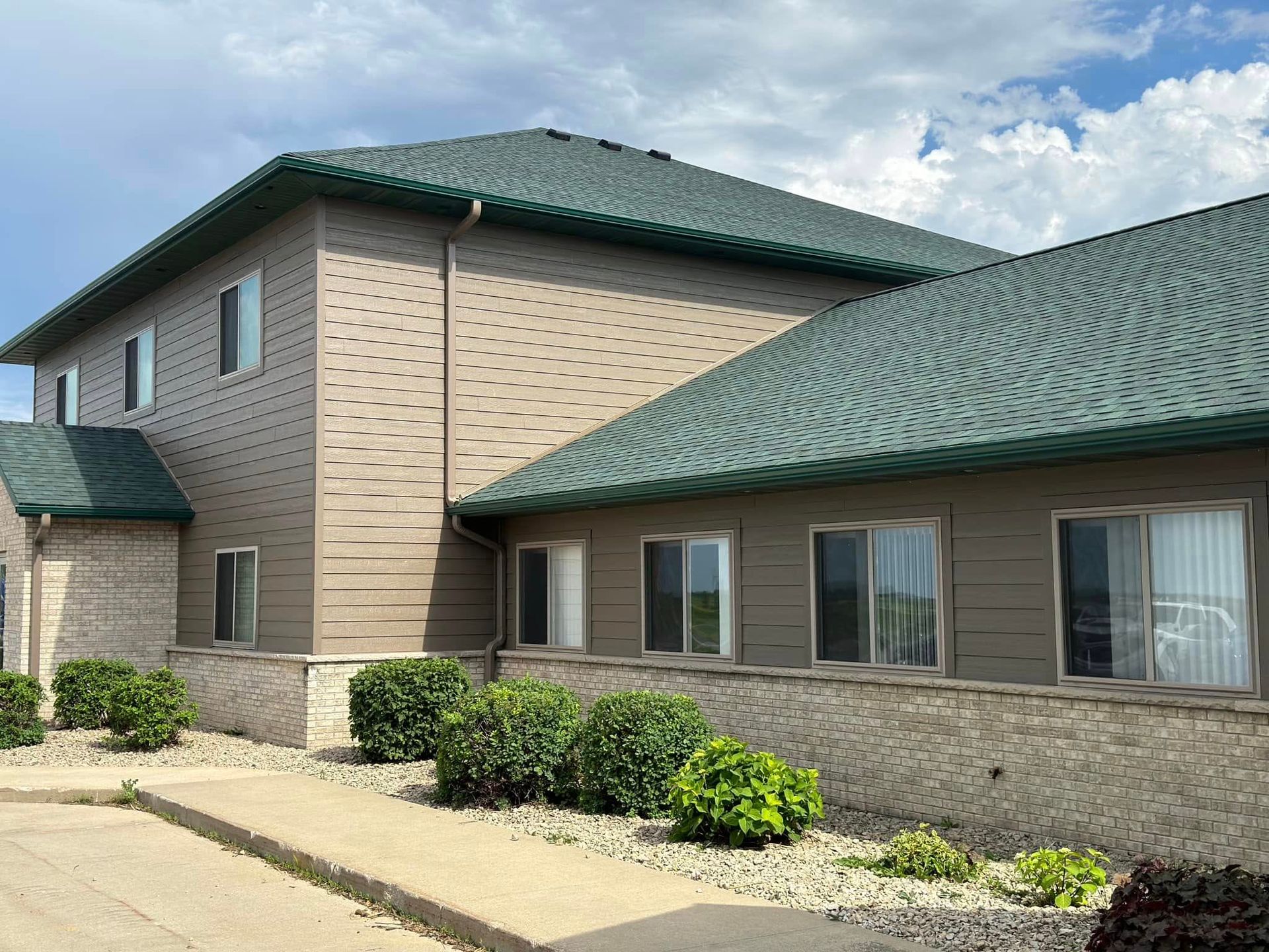 Two-story building with green roof, tan siding, and windows. Bushes line the foundation.