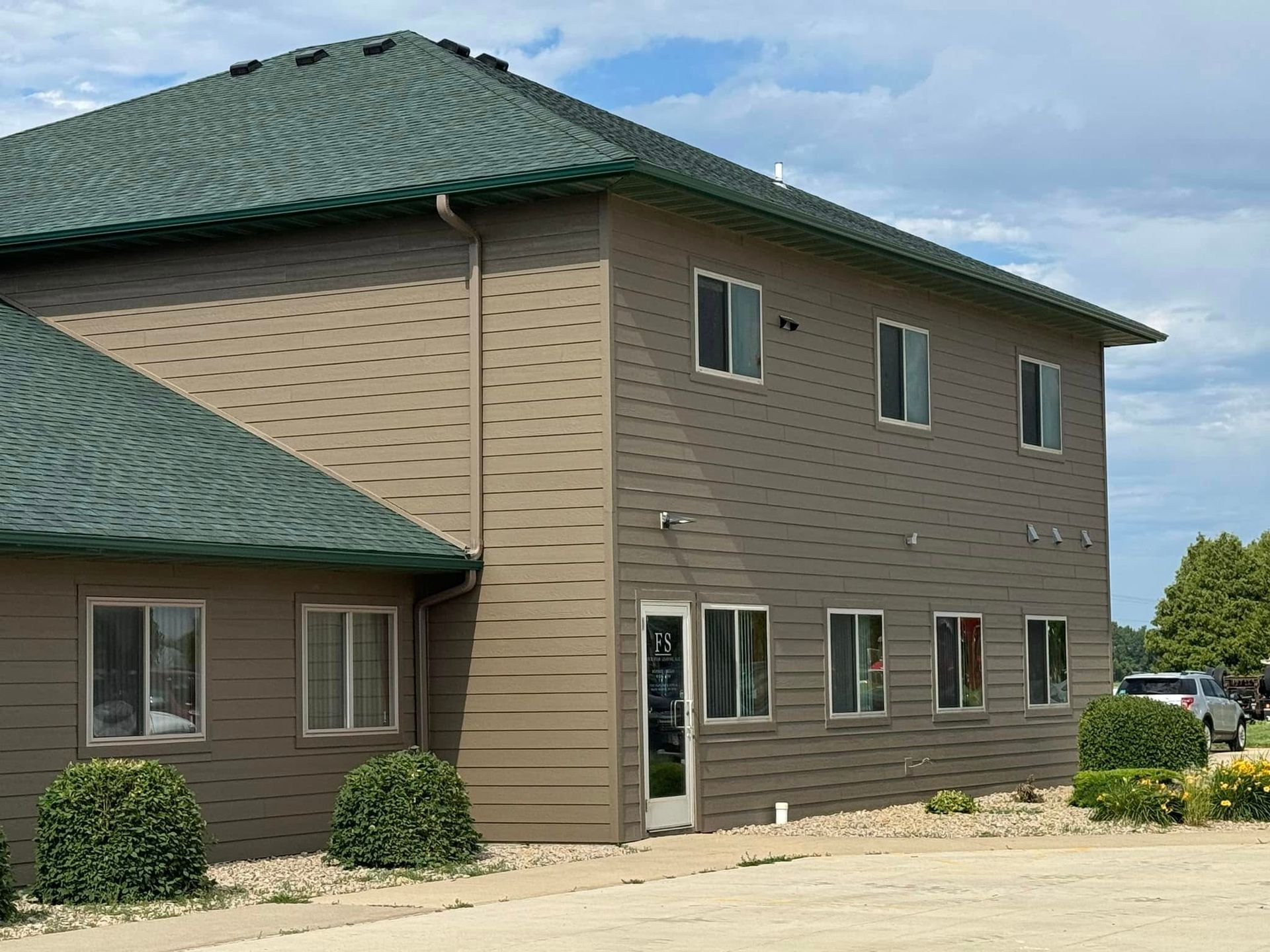 Tan building with green roof, several windows, and landscaping against a cloudy sky.