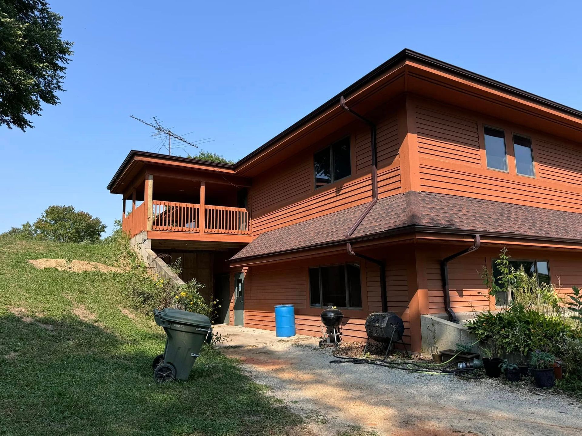 Two-story wood-sided house with balcony and brown shingle roof, on a hillside with blue sky.