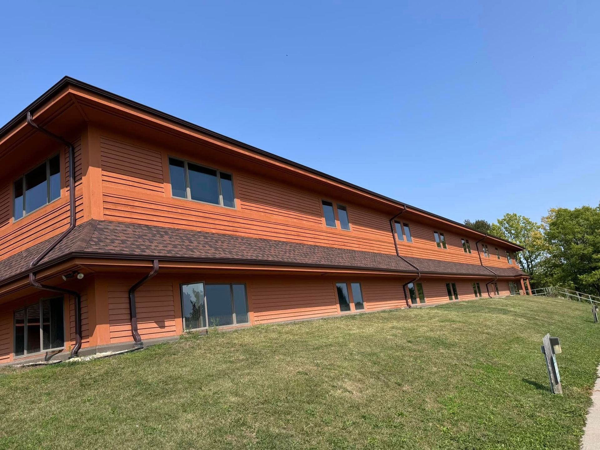 Brown building with many windows and a dark brown roof on a grassy hill under a blue sky.