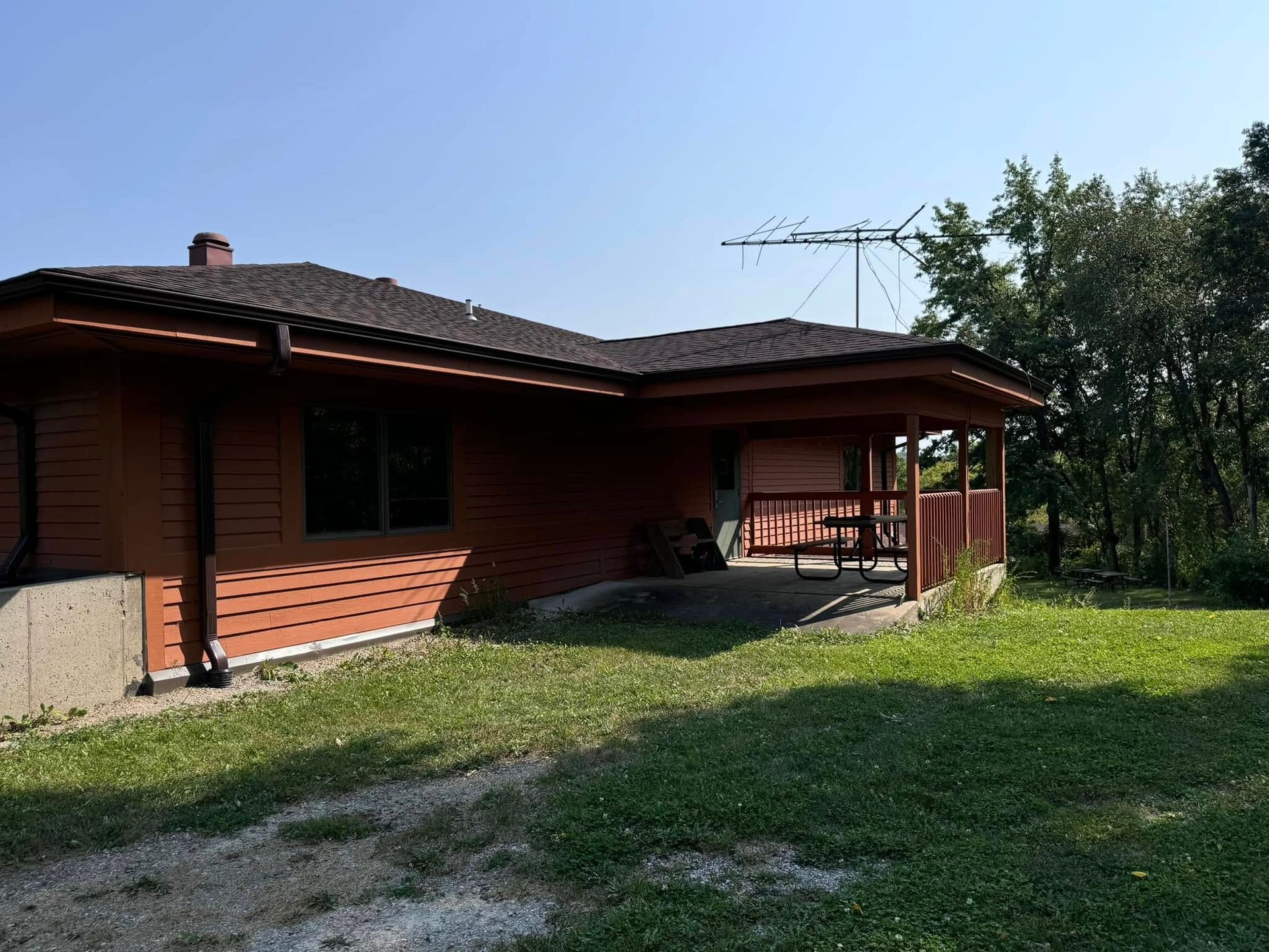 Brown house with porch and antenna on a sunny day. Green grass and trees in the yard.