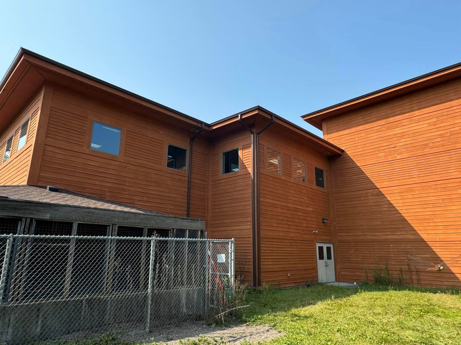 Brown wooden building with several windows. White door. Chain-link fence and grass in foreground.