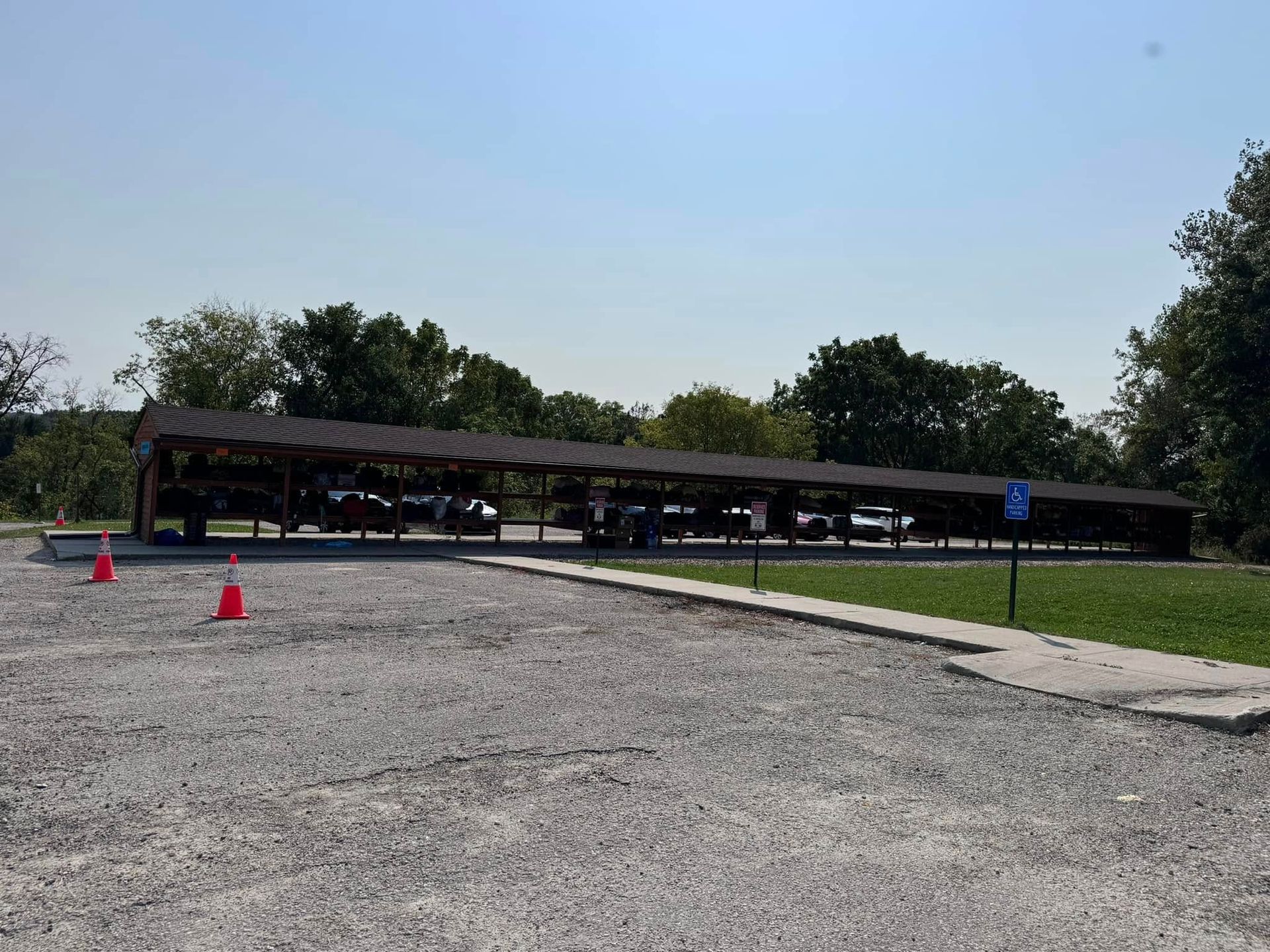 Gravel parking lot with cars parked under a long wooden shelter; blue handicap sign.