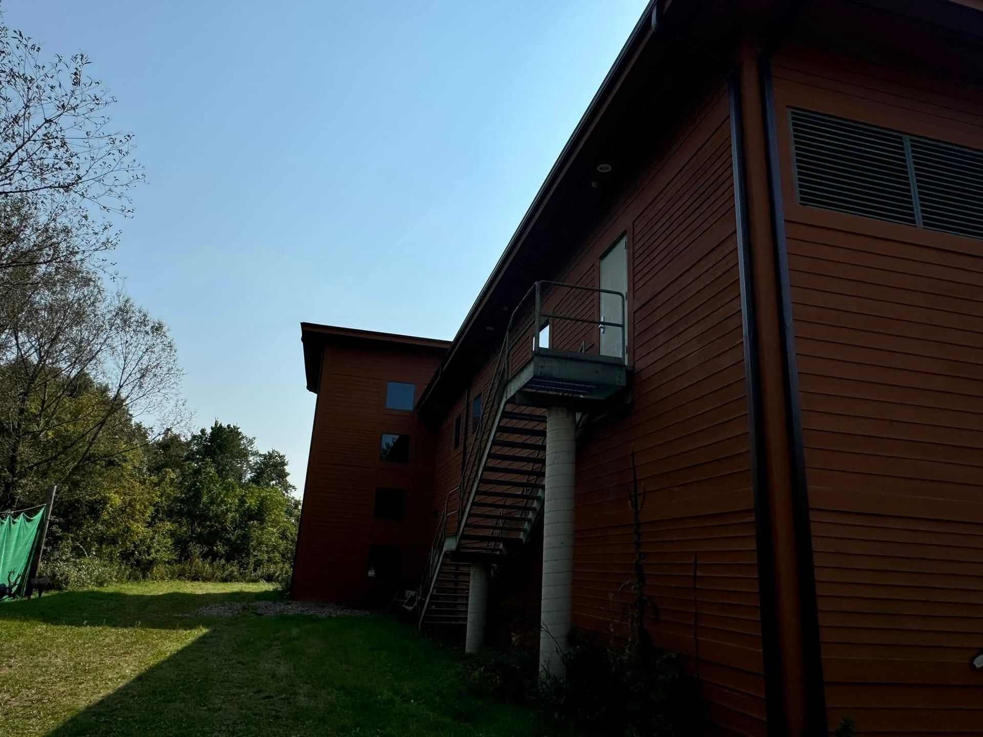 Brown wooden building with exterior staircase; green grass and trees in the background under a blue sky.