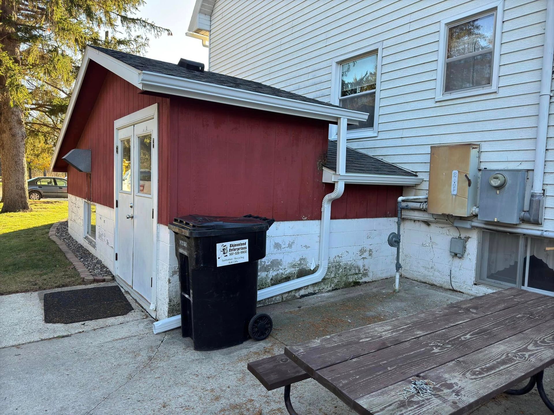 Red shed attached to a white building. Black trash can, picnic table, and grassy area present.
