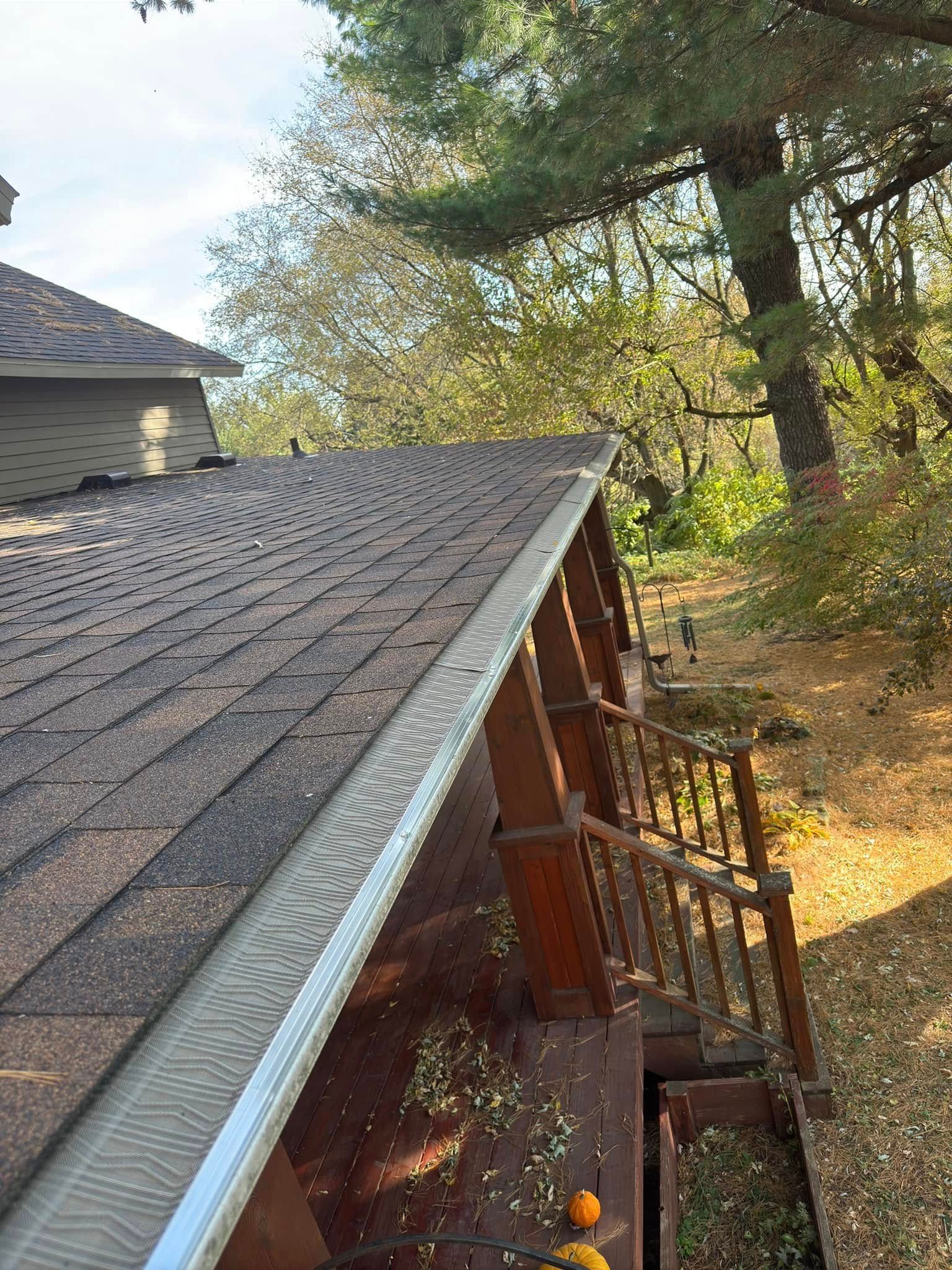 View of a brown shingled roof with a gutter guard, next to a deck with a railing, trees in the background.