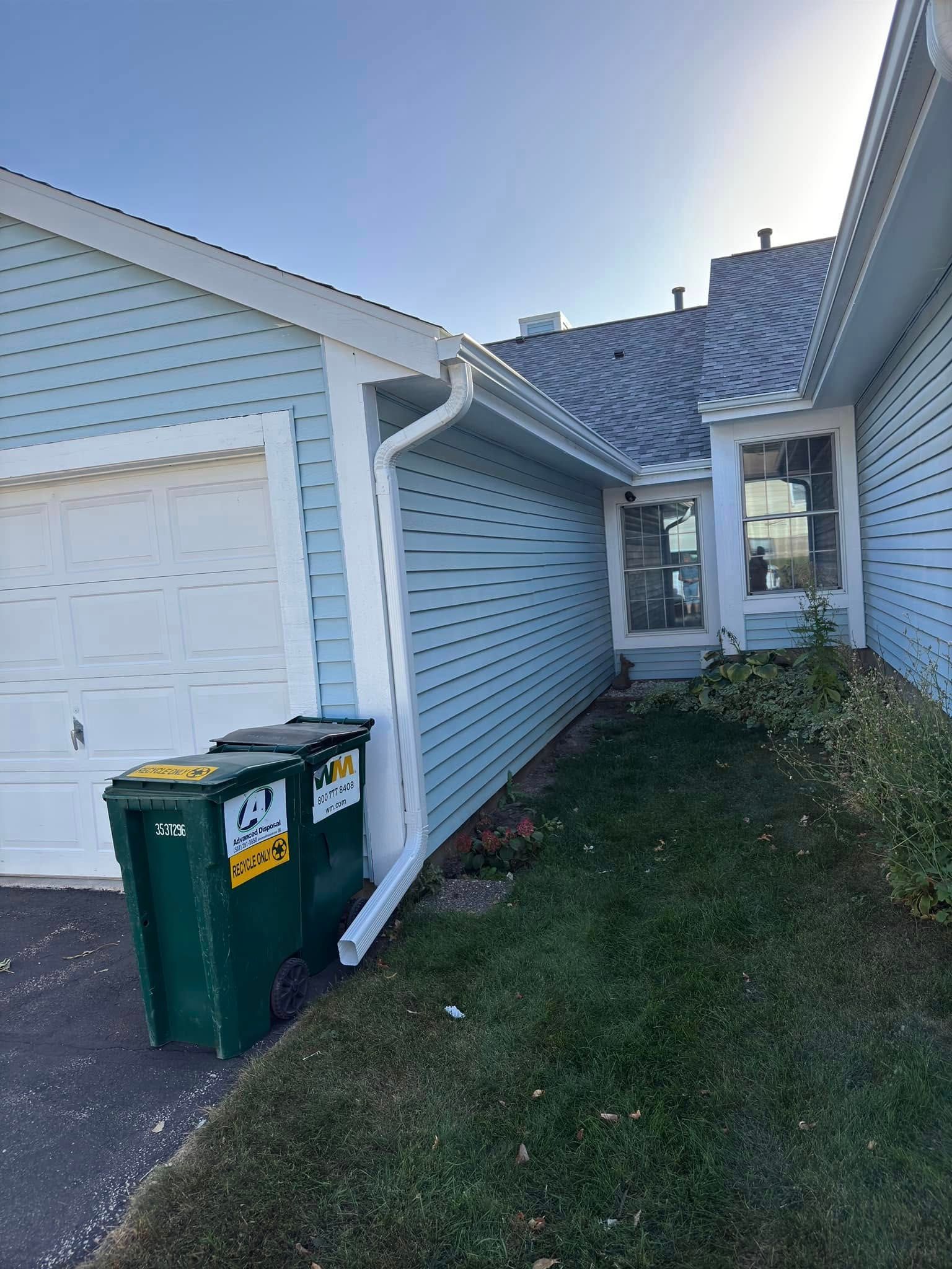 Blue-sided house with a garage and two green trash cans. White trim, blue sky, and a grassy area.