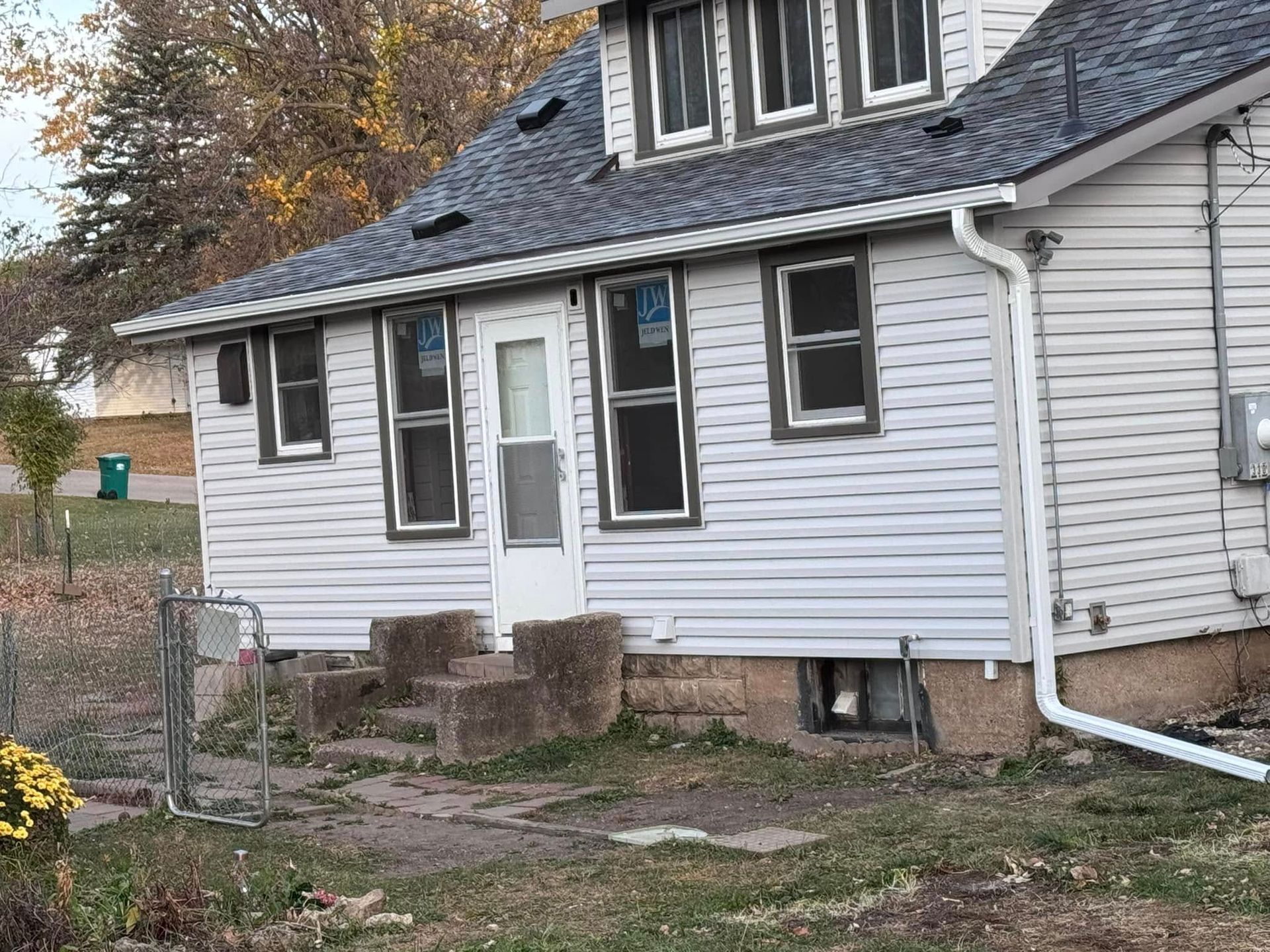 A two-story house with light siding and dark-trimmed windows. A white door is centered, and a concrete path leads up to it.