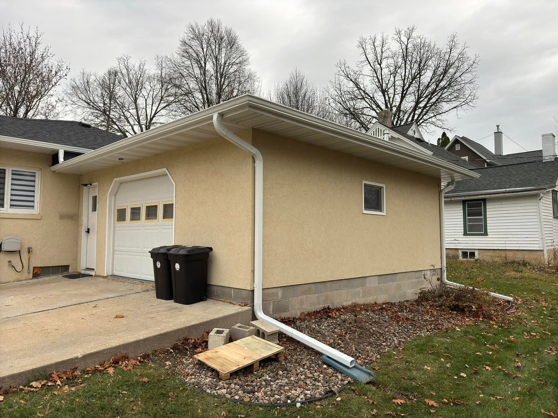 Tan garage with a white garage door, black trash cans, and a concrete driveway.