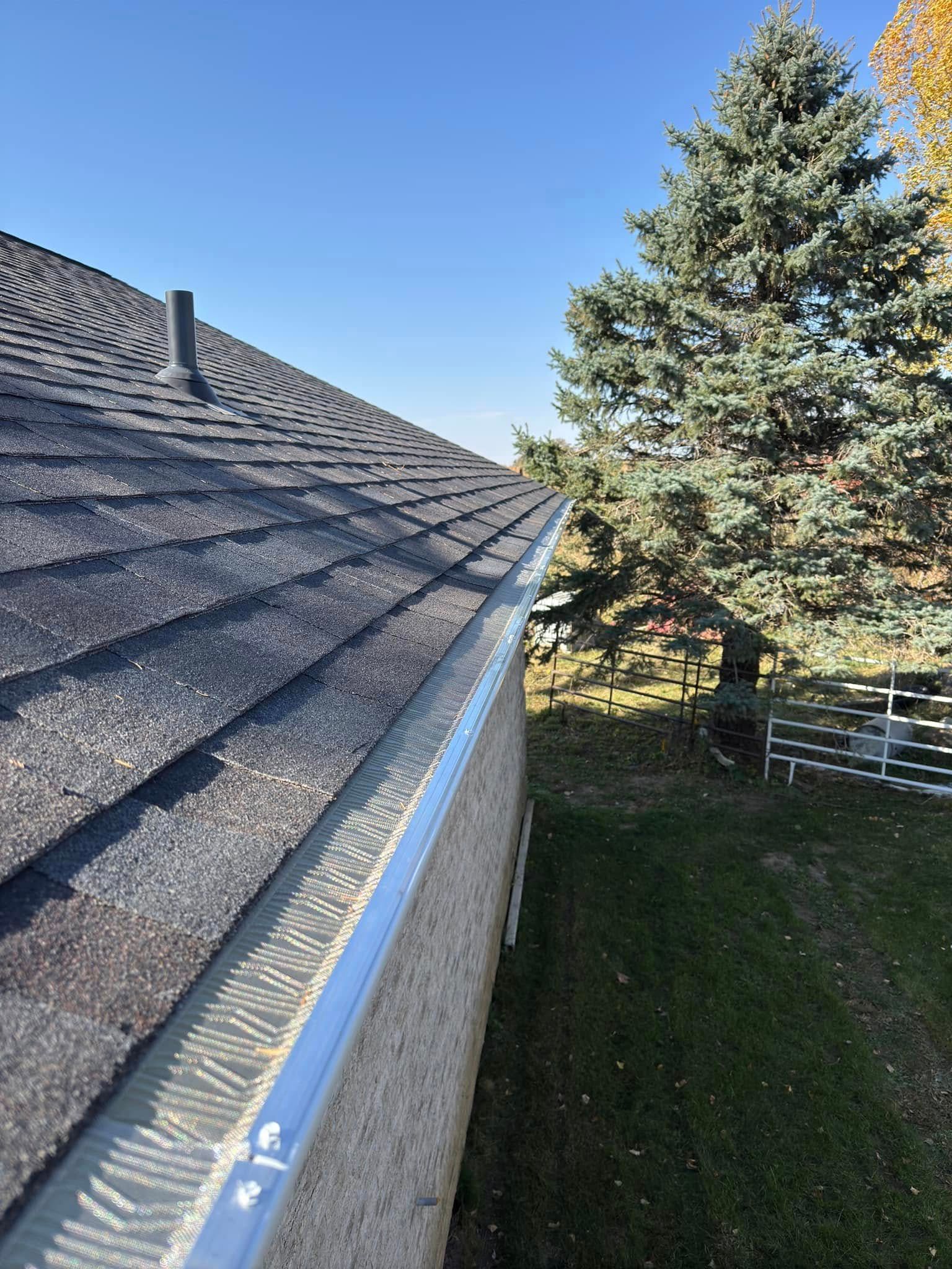 Gutter with leaf guard installed on a house with a dark roof, a tree in the background, and a blue sky.