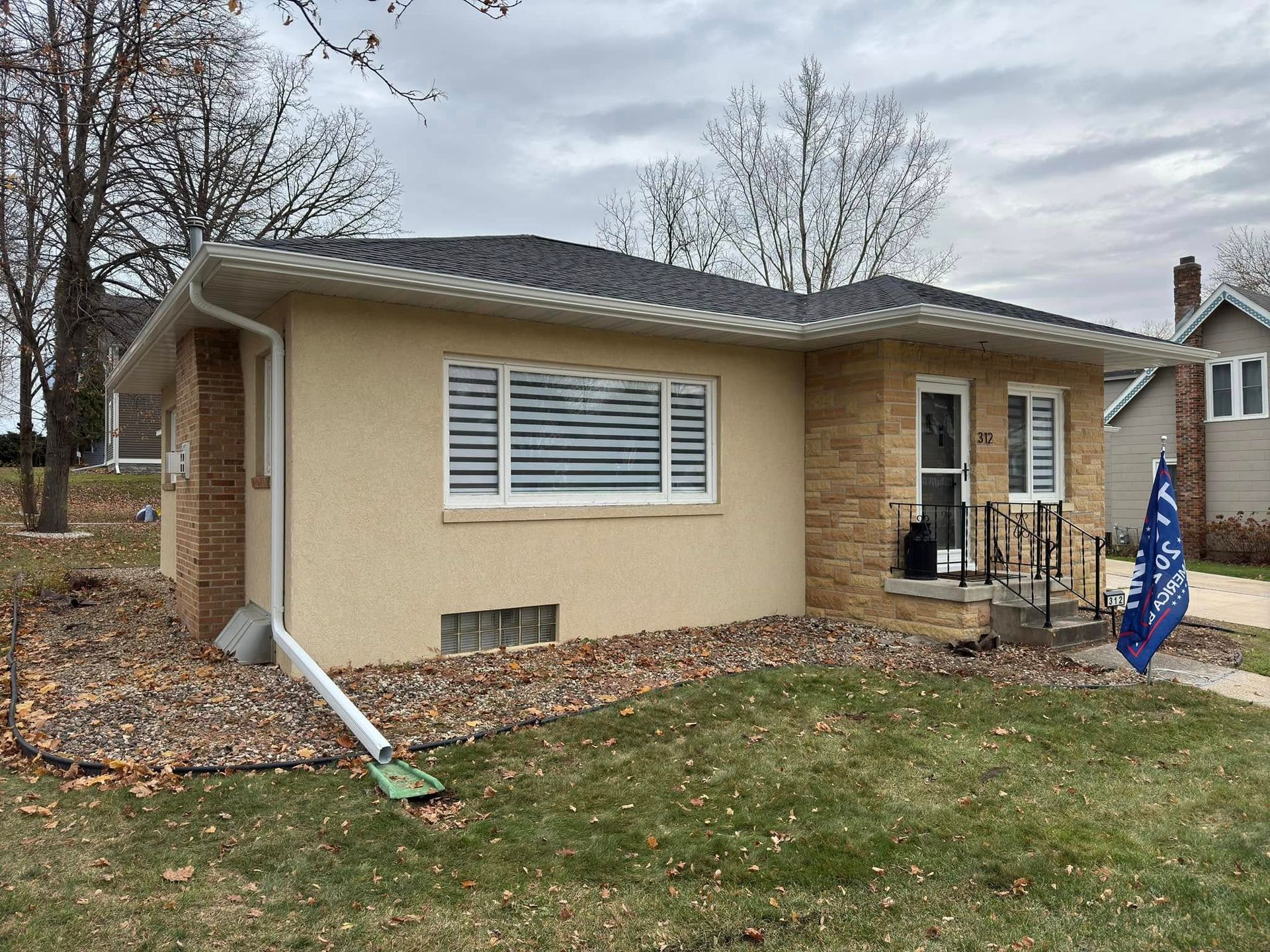 Tan house with brick accents, dark roof, and white shutters on a cloudy day.