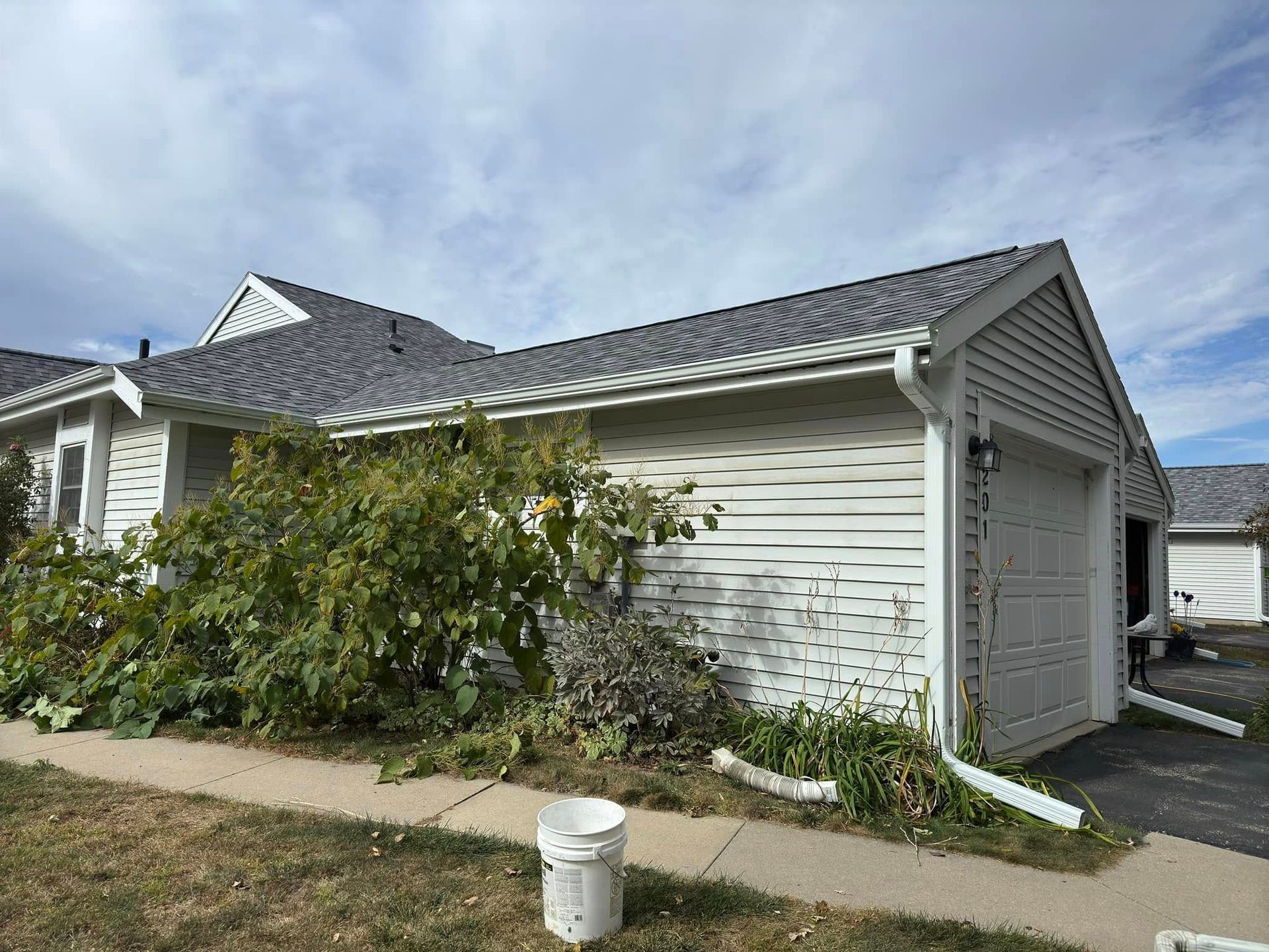 White house with gray roof, garage door, and overgrown bushes in front. Cloudy sky.