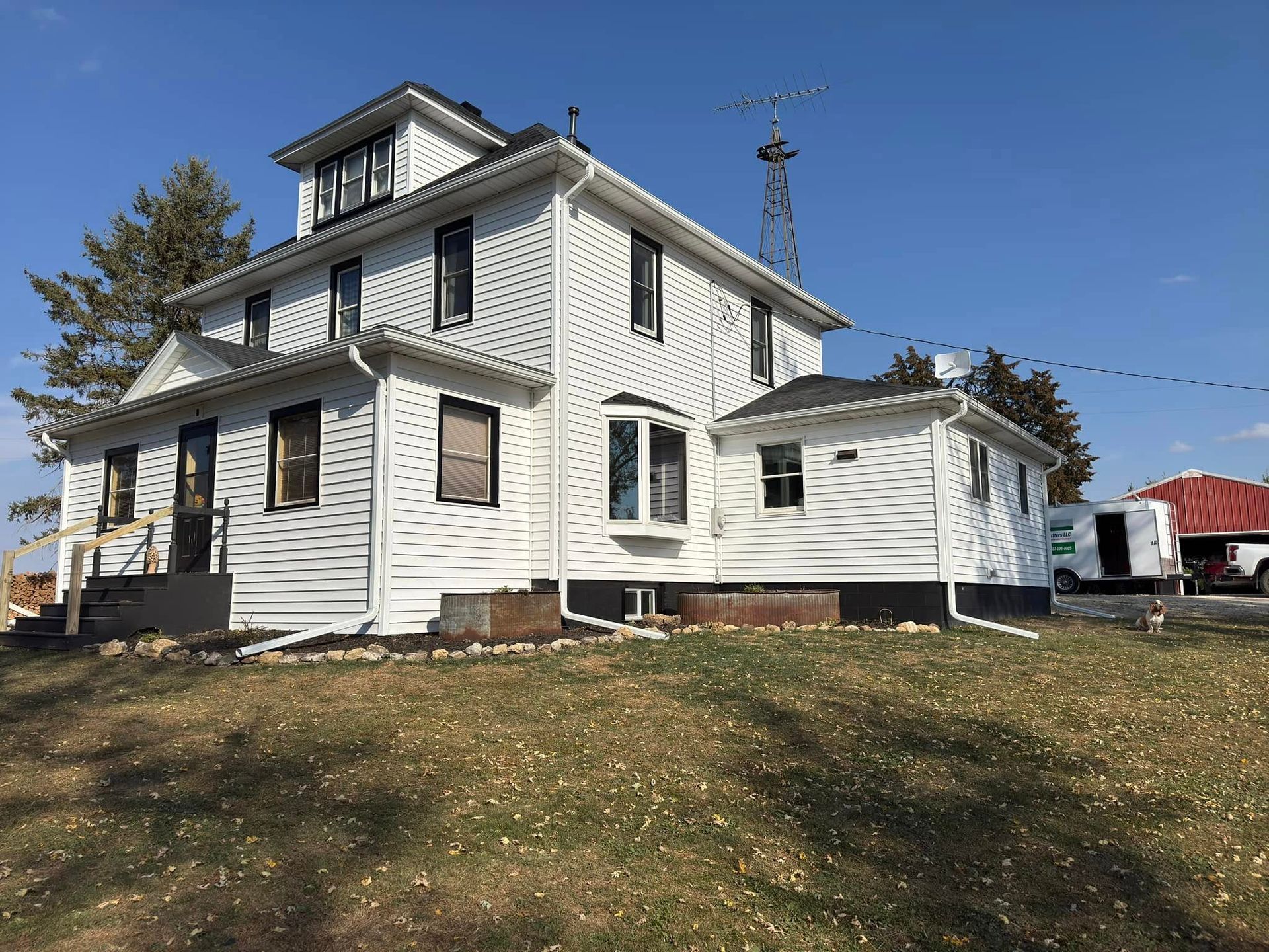White two-story house with black trim, bay window, and a windmill in the background on a sunny day.