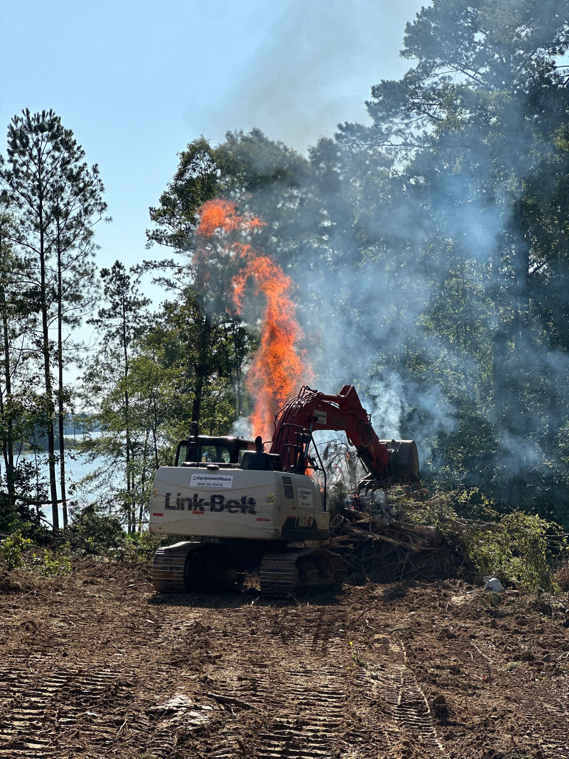 Excavator burning debris in a wooded area near water, flames and smoke rising.