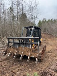Excavator burning brush in a wooded area; smoke and flames visible, trees and lake in the background.