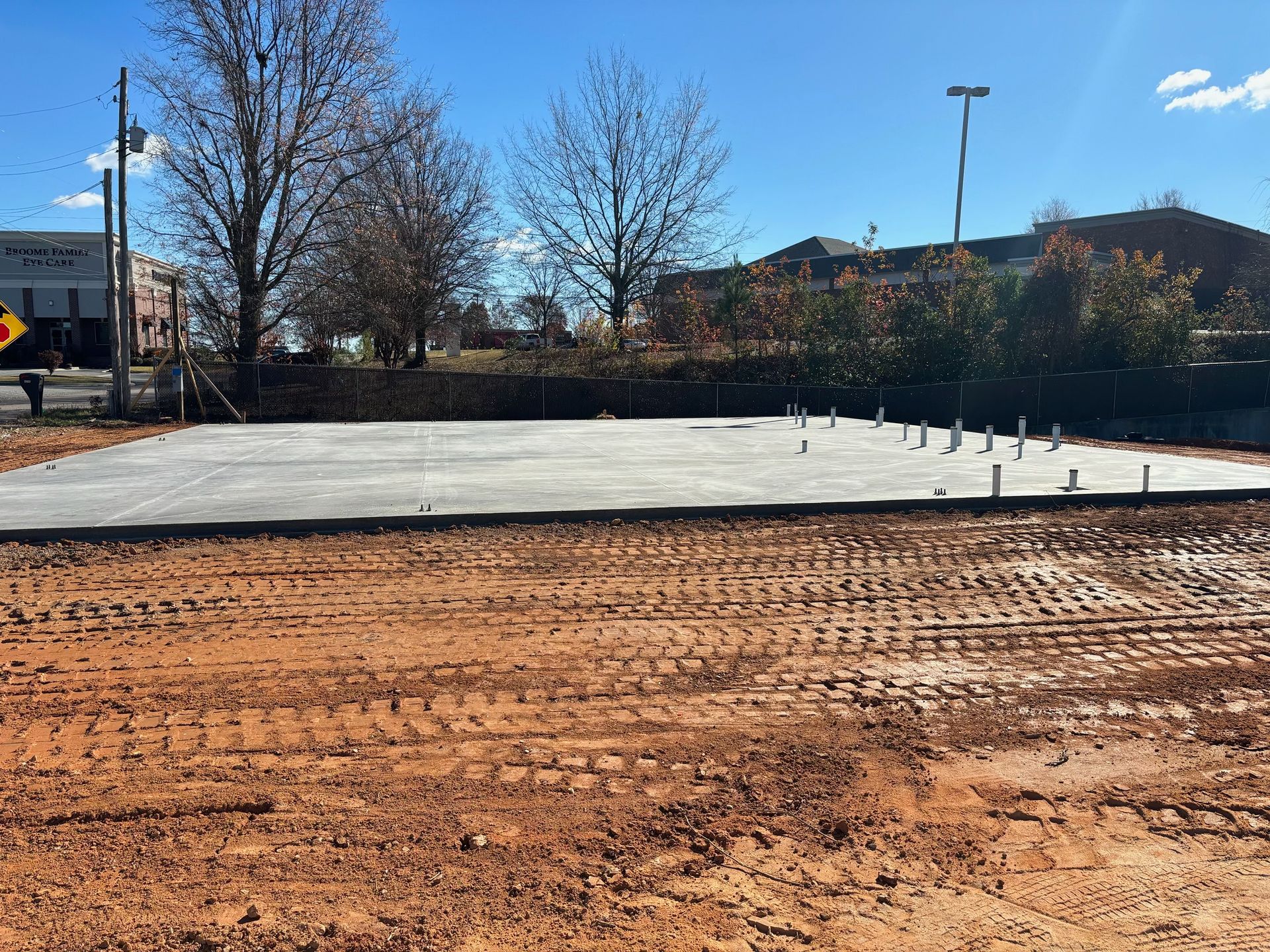 Construction site with a concrete slab and exposed pipes; dirt in foreground, trees and buildings in background.
