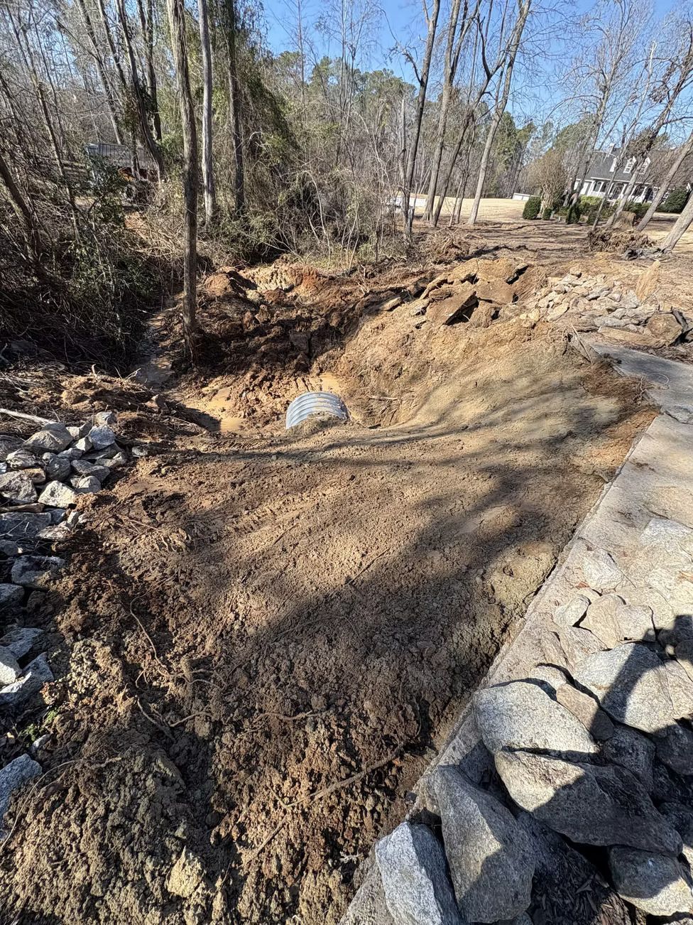 Dirt-filled stream bed with a culvert, surrounded by rocks, bare trees, and a sunny sky.