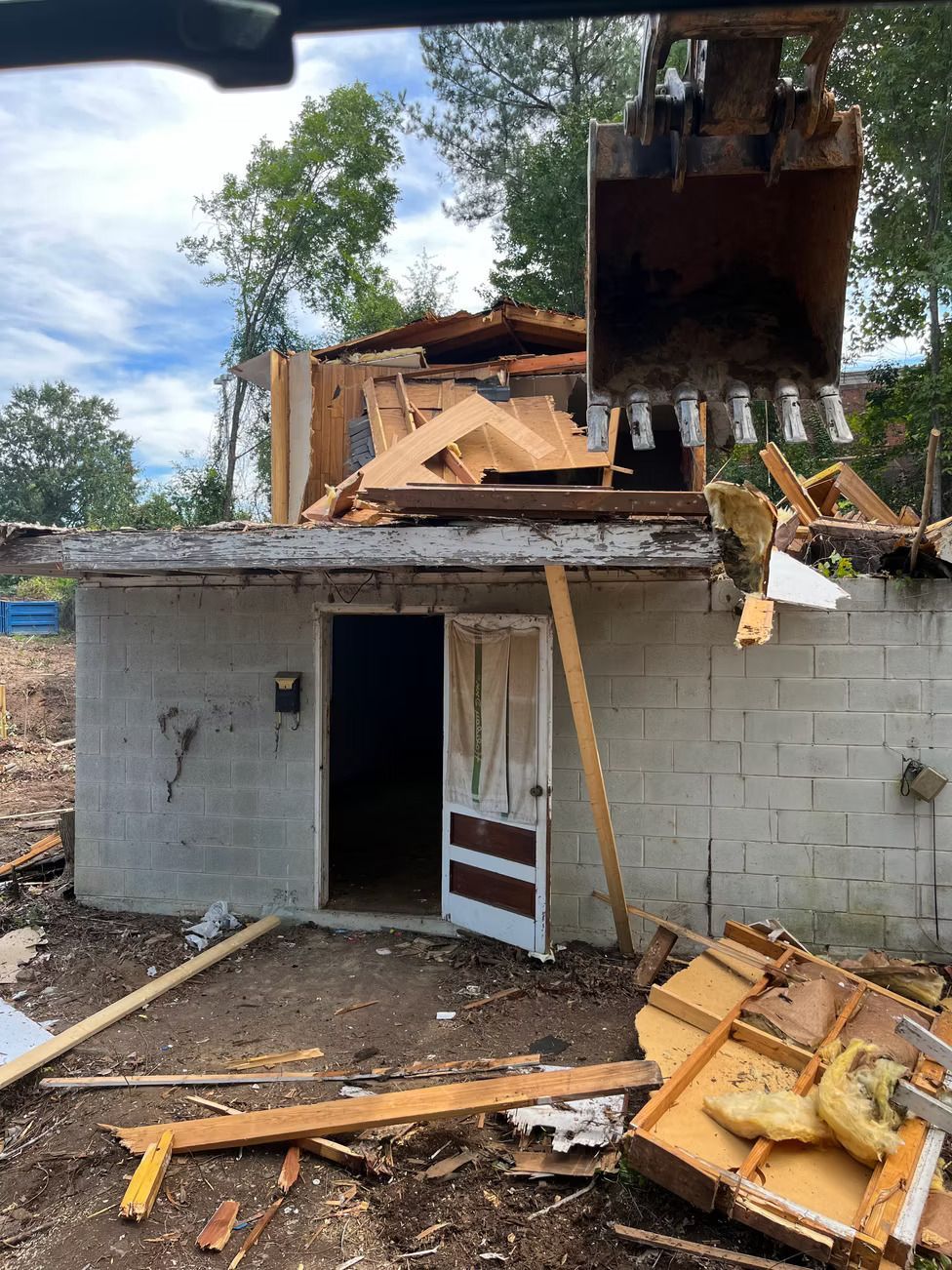 An excavator demolishes a small, cinderblock building with an open doorway; debris scattered on the ground.