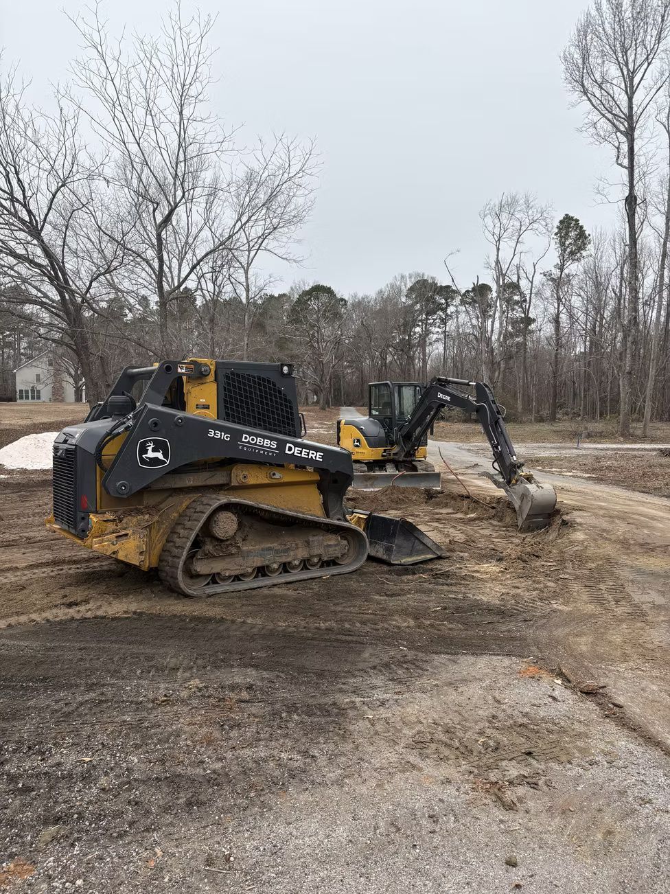 Two yellow construction vehicles on a dirt road near trees. One is a tracked skid steer, the other is an excavator.