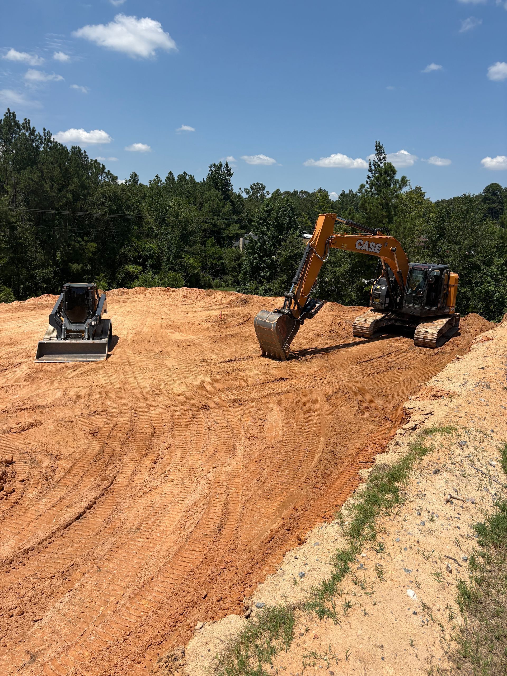 Excavator and concrete mixer on a red-dirt construction site, with trees and blue sky in background.