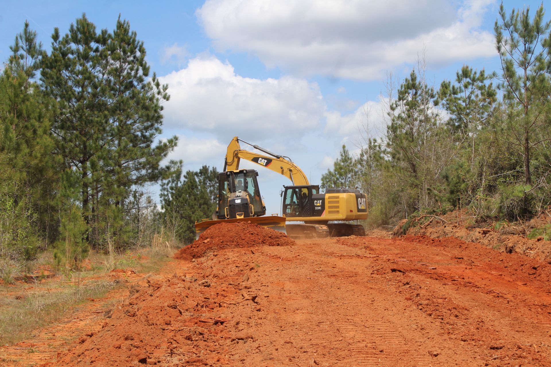 Excavator and concrete mixer on a red-dirt construction site, with trees and blue sky in background.