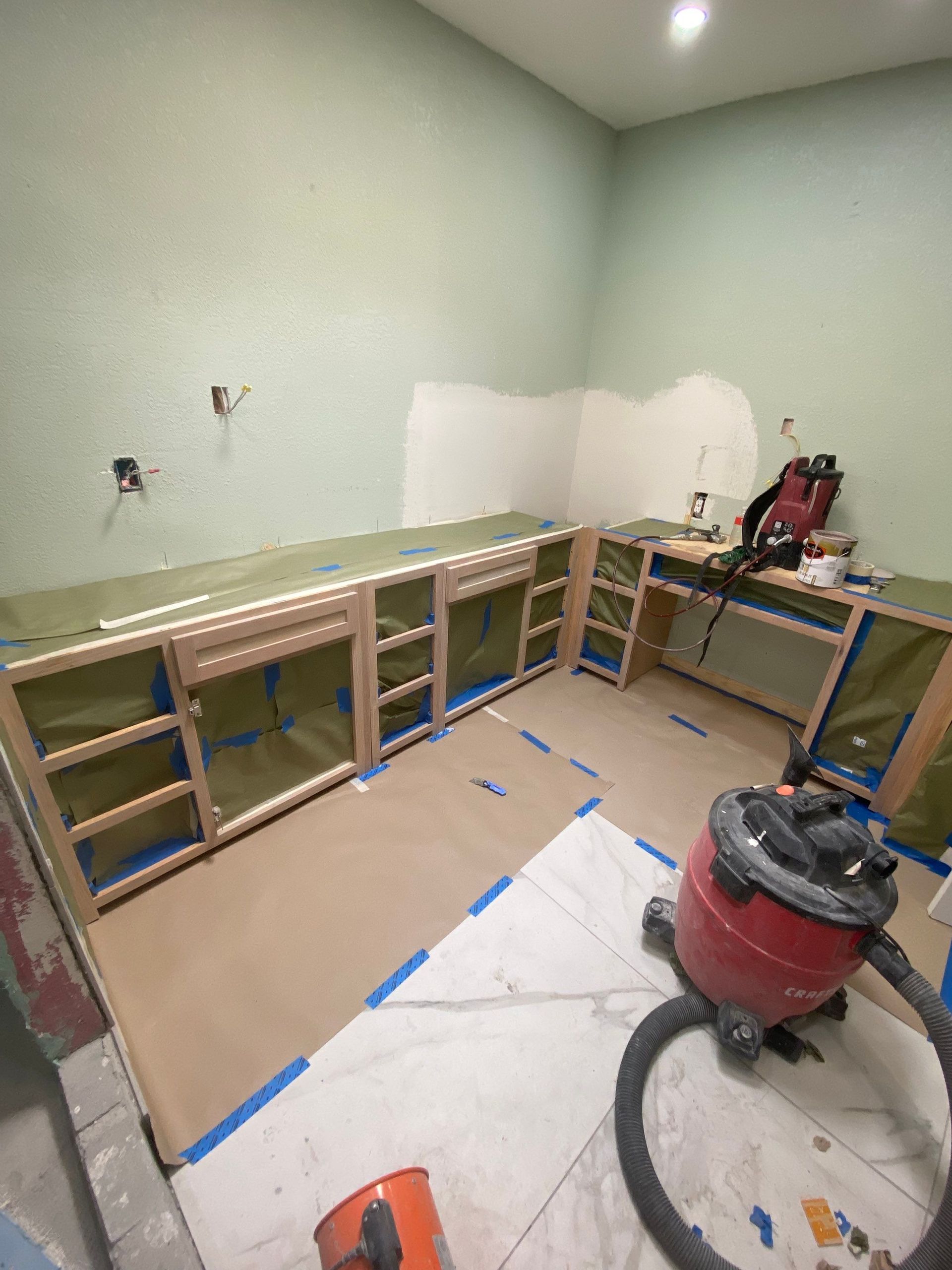 Cabinets under construction in a room, covered with protective materials. A vacuum cleaner sits on the floor.