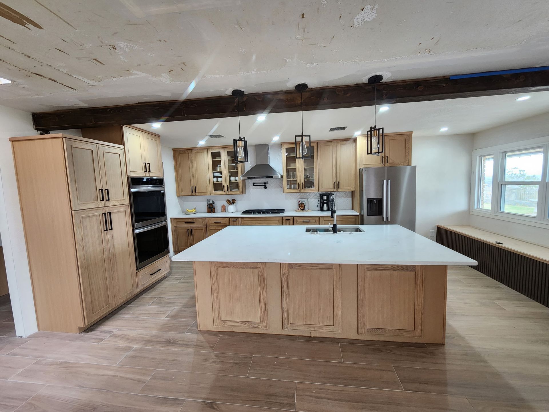 Modern kitchen with light wood cabinets, white countertops, and an island. Stainless steel appliances and exposed beams.