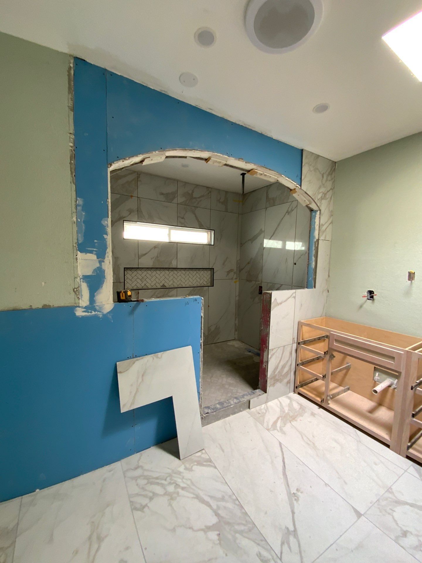 Bathroom under construction, blue wallboard, archway to tiled shower, bare wood cabinet, light-colored tile floor.