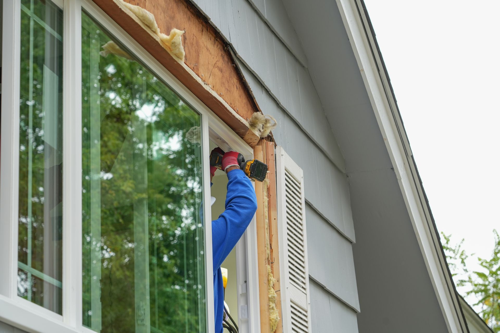 Person installing a window, working on exterior siding of a gray house.
