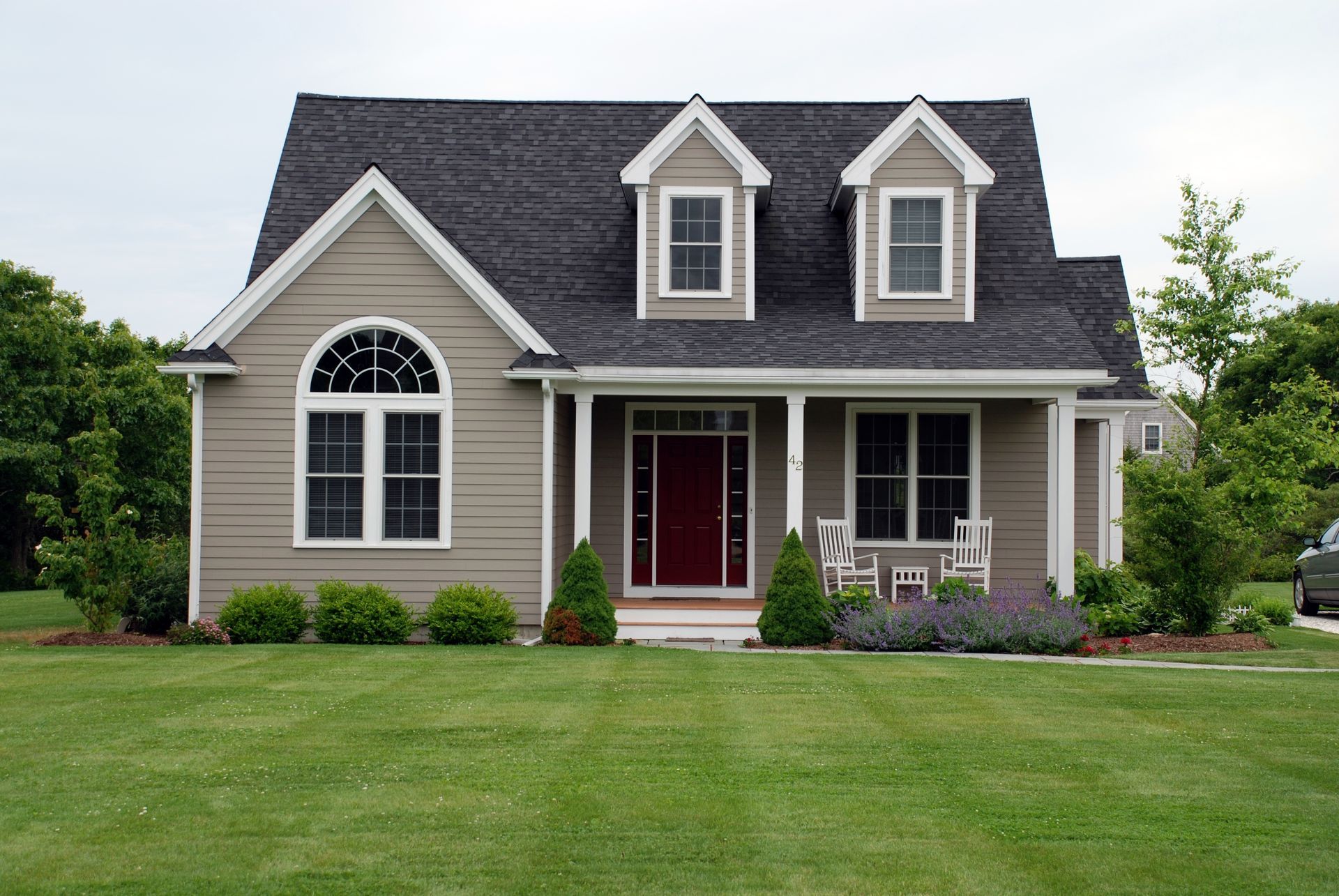 Tan house with dark roof, arched window, red door, porch, and dormers on a green lawn.