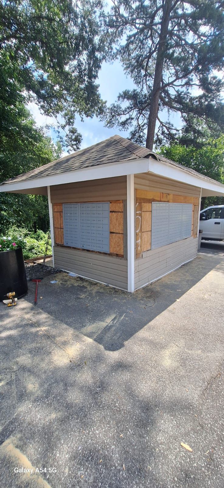 Small wooden kiosk with gray siding and roof, in a gravel parking area, surrounded by trees.