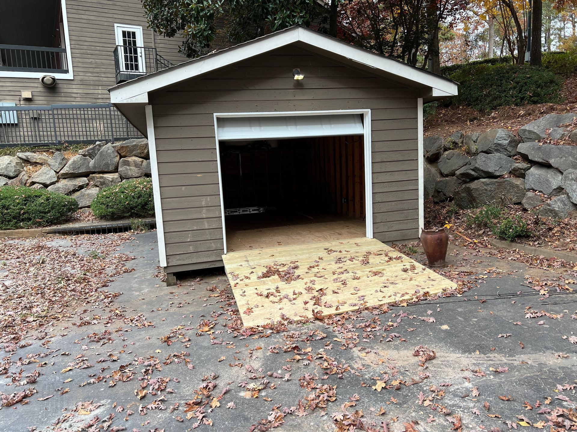 Brown shed with an open garage door and a wooden ramp on a driveway covered in fall leaves.