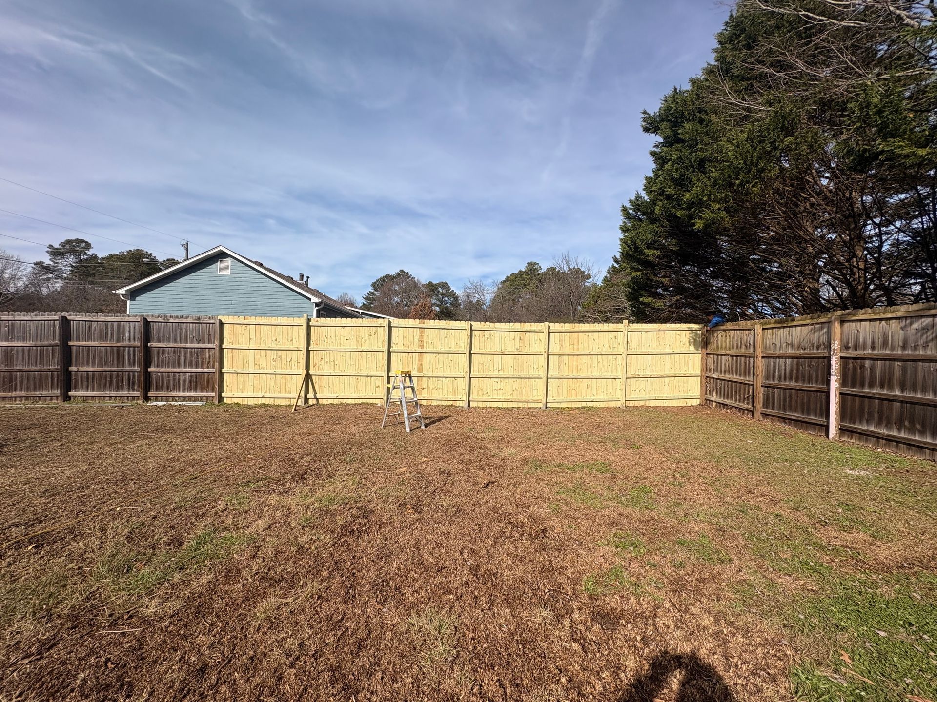 Wooden fence in a backyard, with a light-colored central section and two darker sides. Autumnal yard and a blue house.