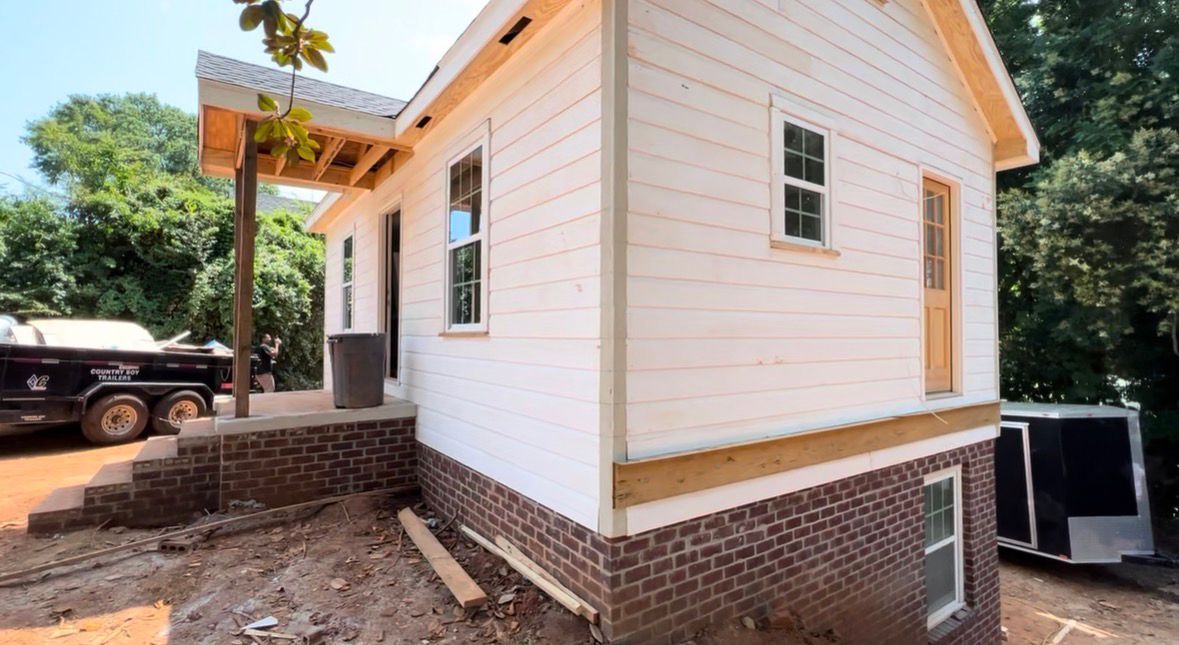 White-sided house under construction with brick foundation, wooden porch, and a truck parked to the left.