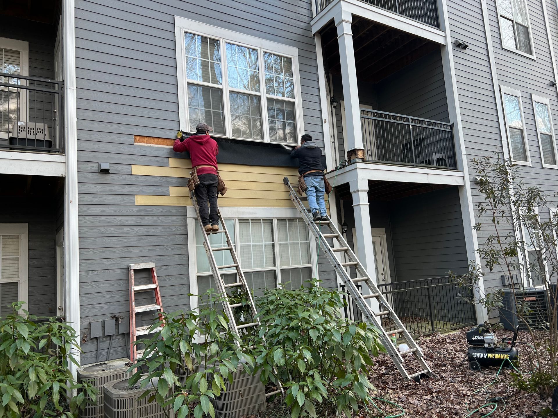 Two workers on ladders repair siding on a building with gray siding and white windows.