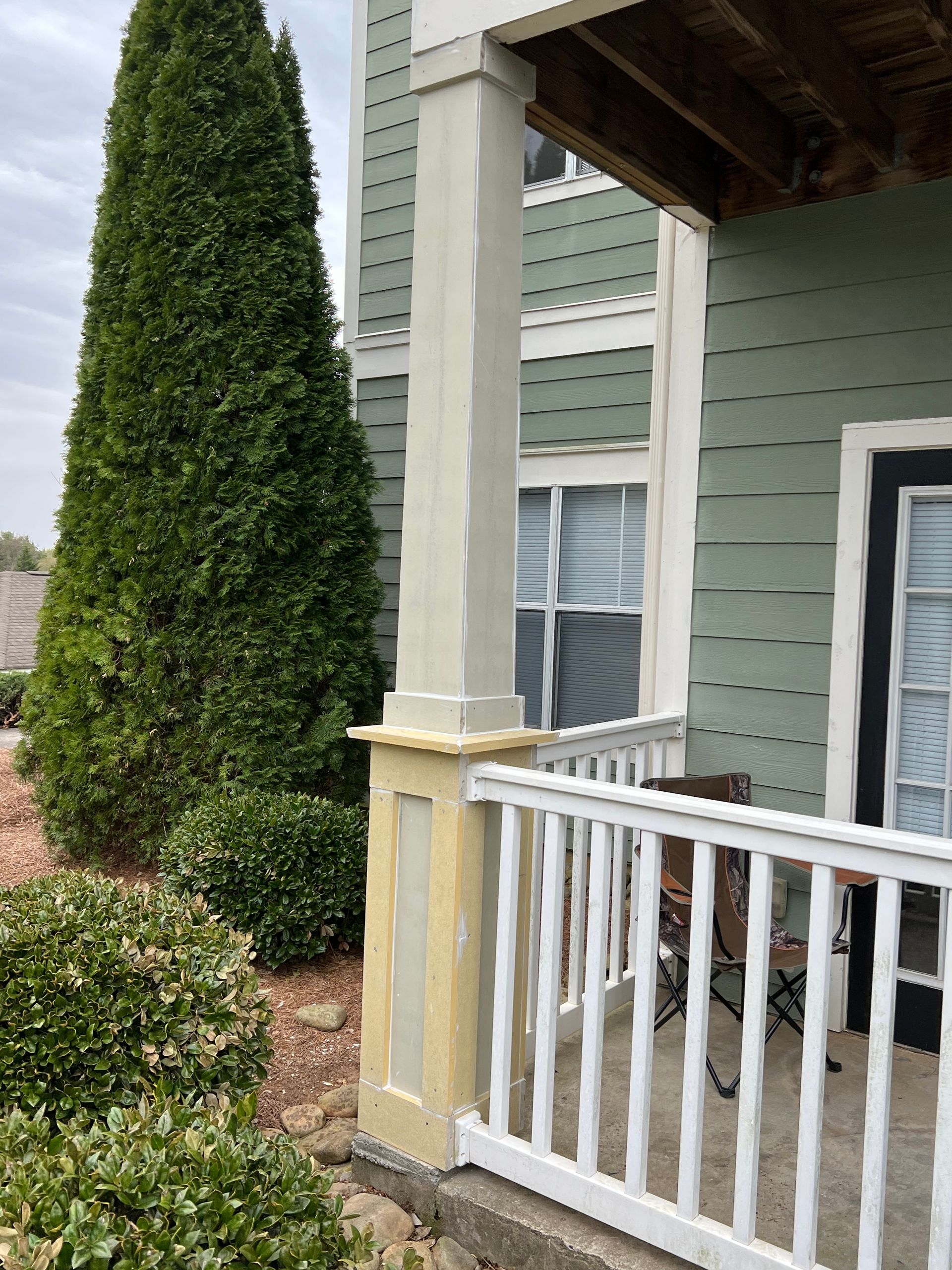 Porch with white railing and light tan pillar next to a green-sided building and evergreen trees.
