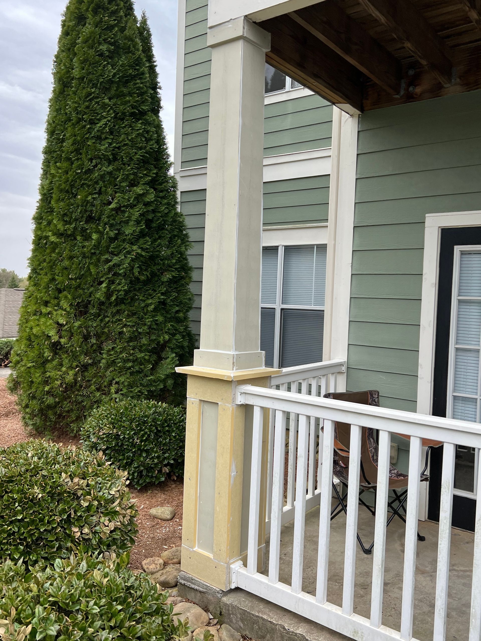 A porch with white railing and a column. Green siding and a tall evergreen tree are visible.