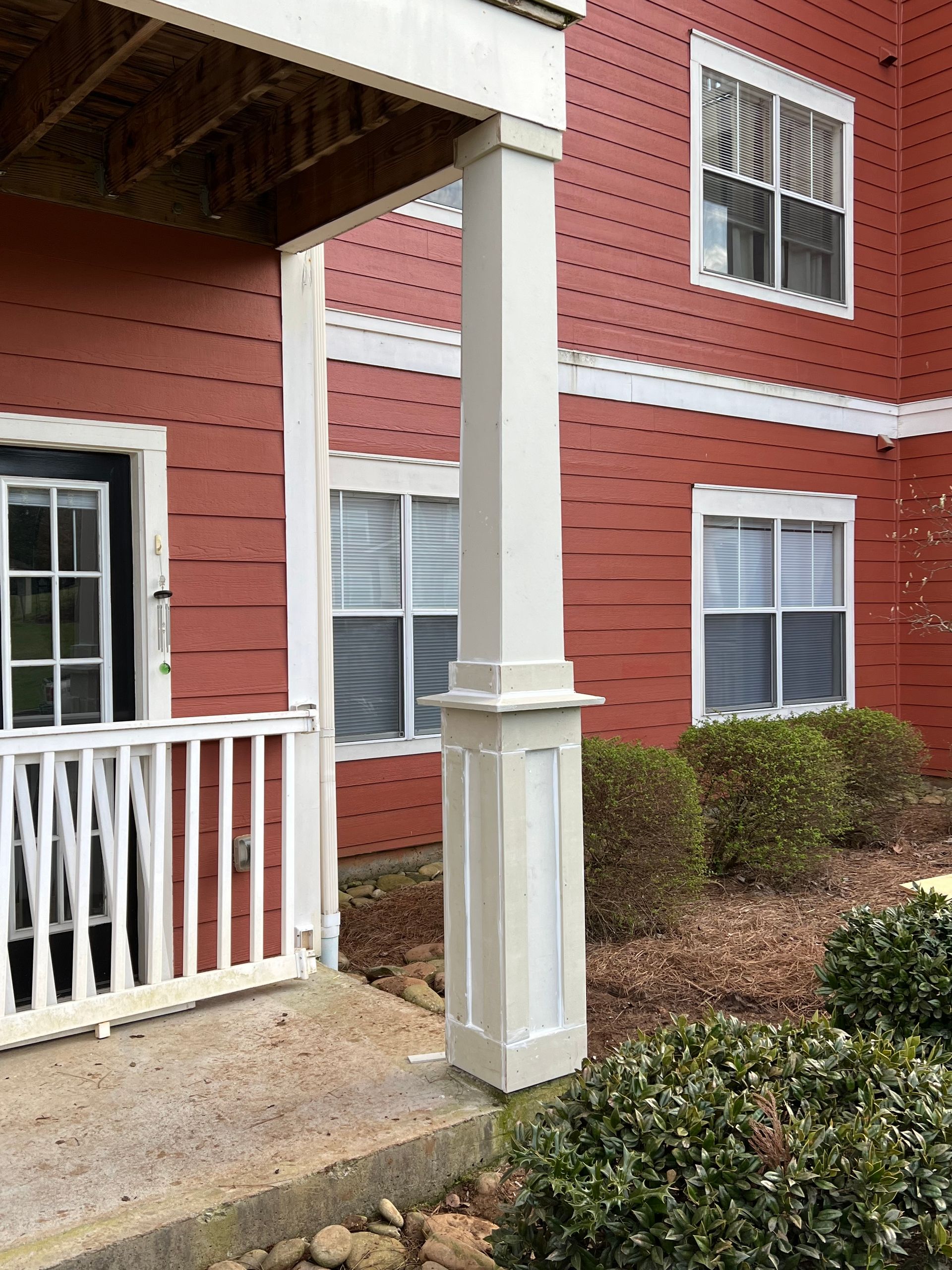 A light-colored column on a building's porch, set against a red exterior with windows and shrubbery.