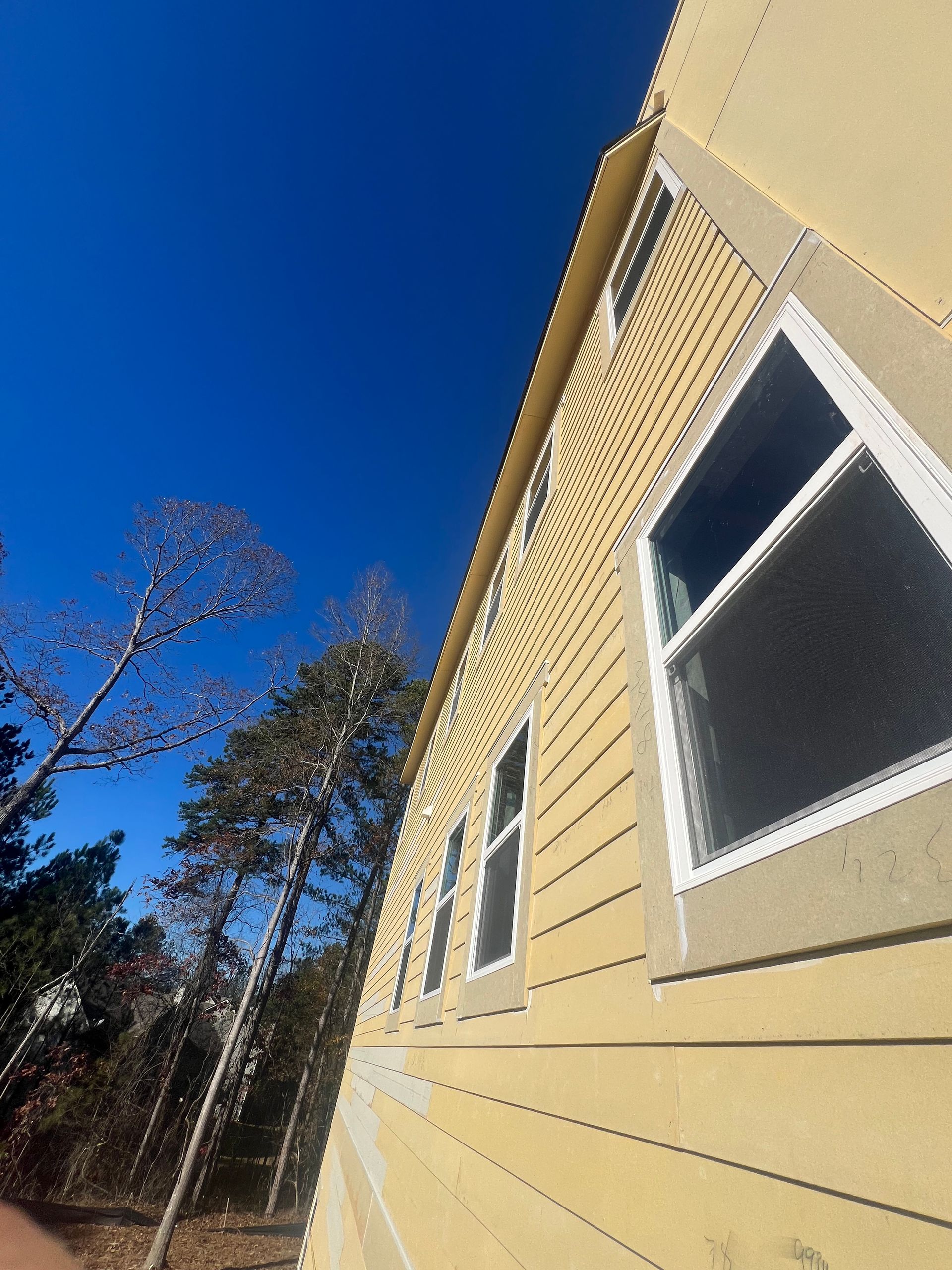 Exterior of a yellow house with white-framed windows against a clear blue sky, trees in the background.