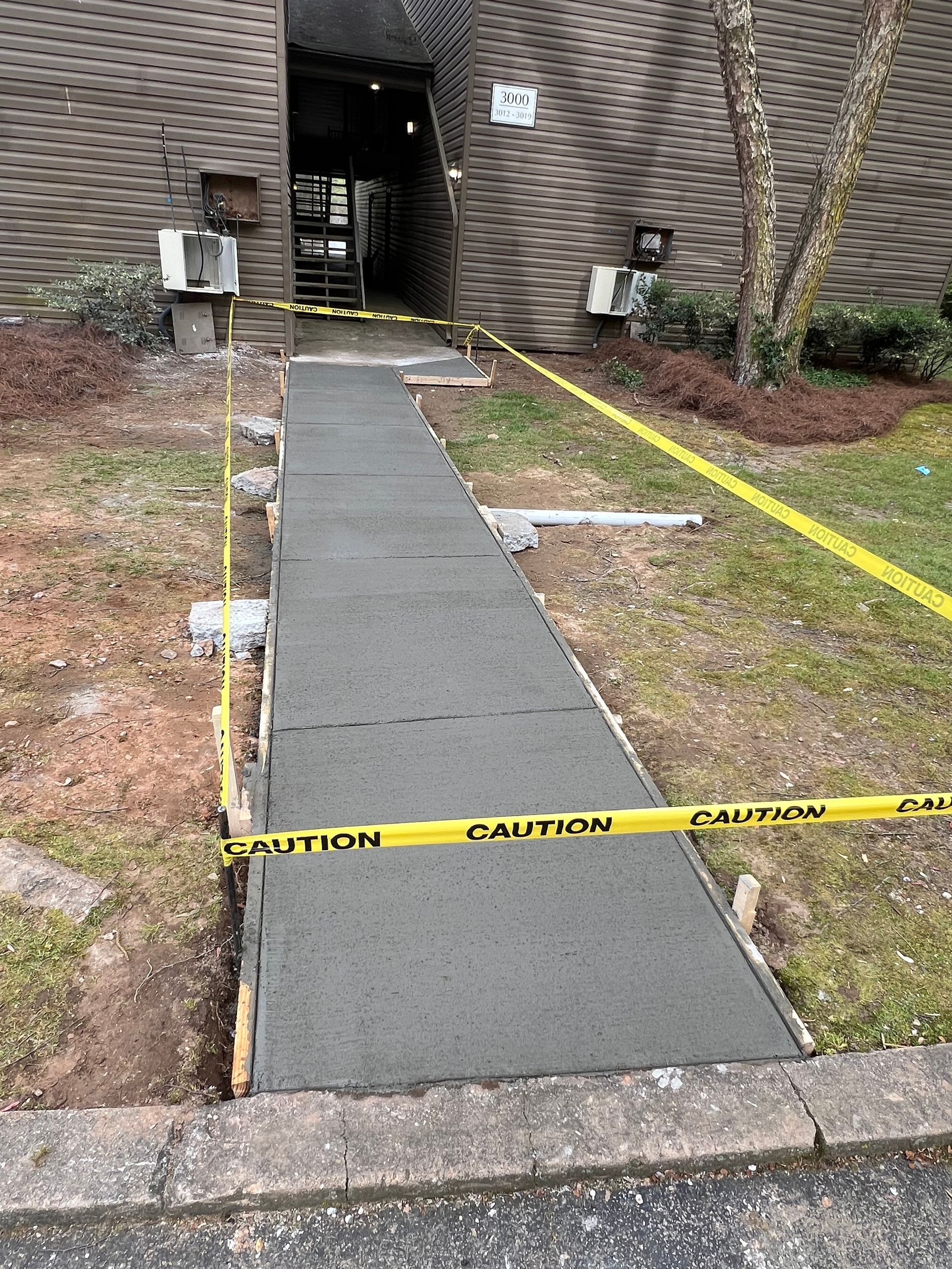 Newly poured concrete sidewalk with caution tape, leading to a building entrance.