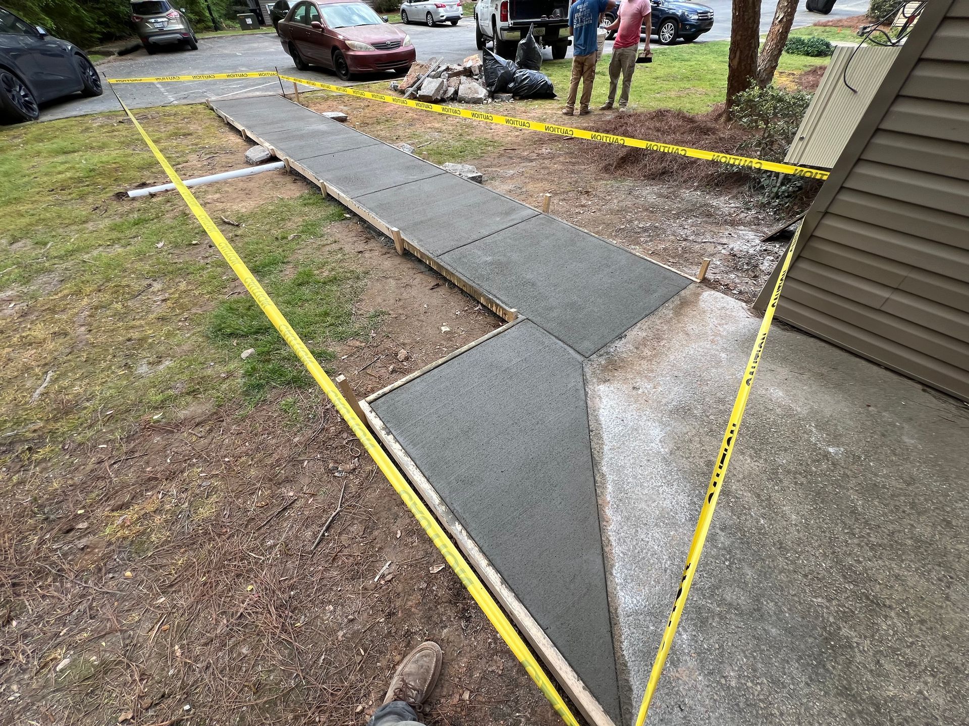 Newly poured concrete sidewalk with wooden forms and caution tape, near a driveway and building.