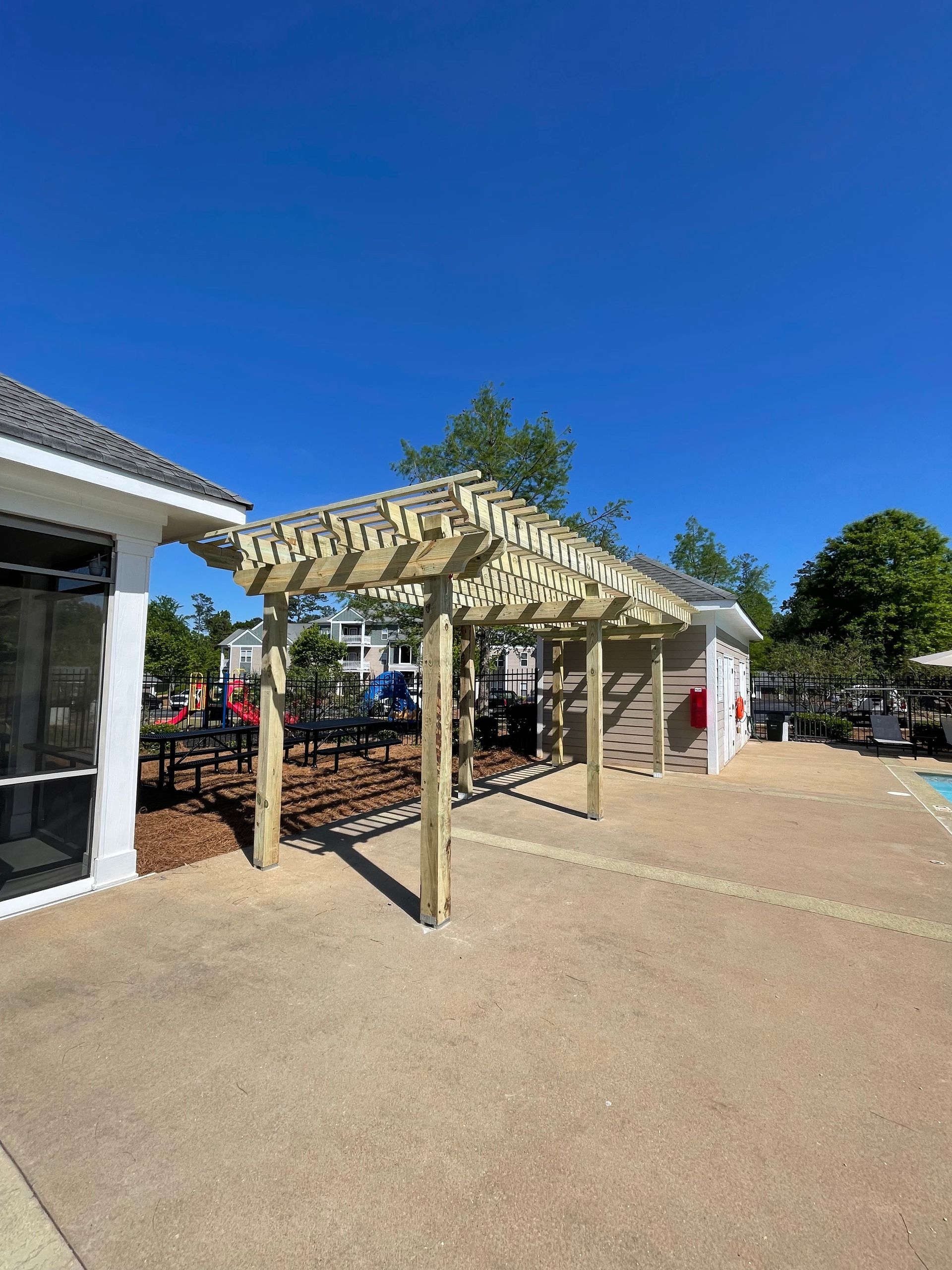 Pergola on a concrete patio next to a building and a pool. Blue sky overhead.