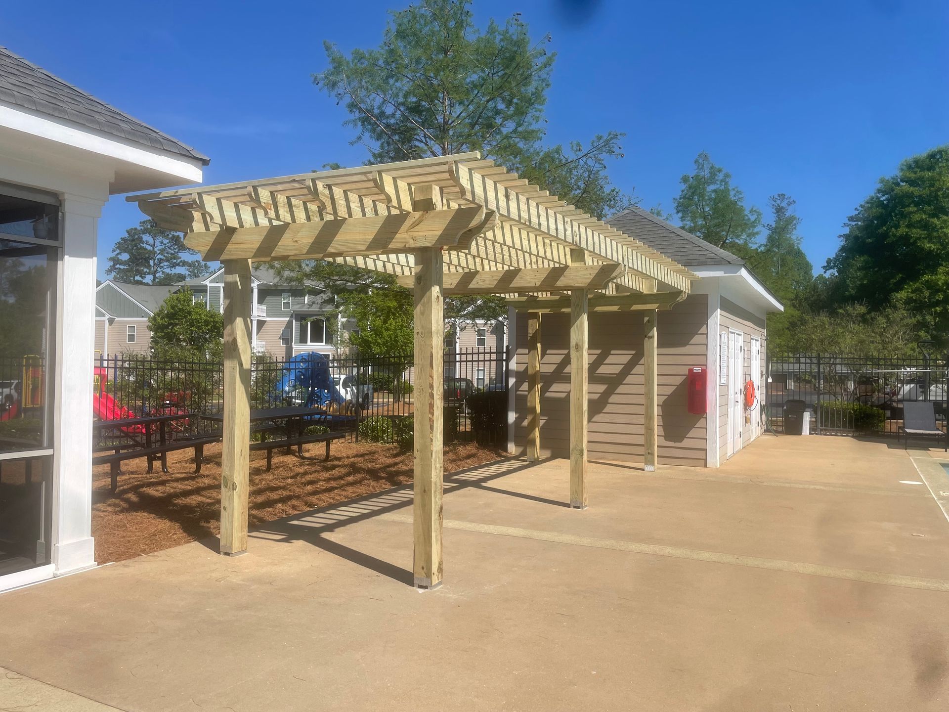 Wooden pergola providing shade over a concrete area beside a building, with a clear blue sky.
